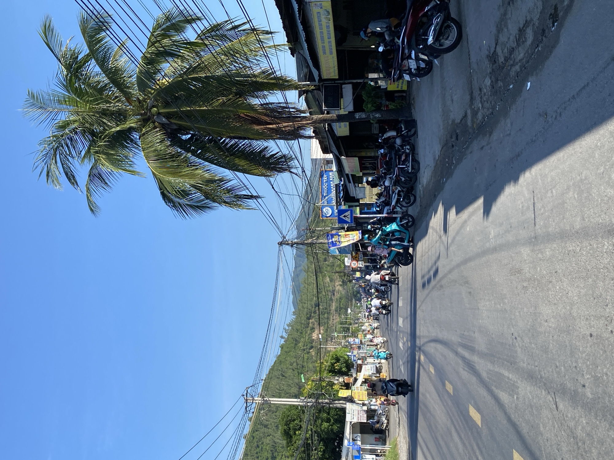 The image depicts a busy street scene in a tropical or subtropical location, possibly Southeast Asia, under a clear blue sky. The road is paved and shows tire marks, with yellow lane markings visible. A variety of motorbikes are being ridden by people wearing helmets, indicating a common mode of transportation. On the right side of the street, there are low-rise buildings with corrugated metal roofs, suggesting commercial establishments or shops. A tall palm tree with dense fronds stands prominently near the buildings, with electrical wires strung above. On the left side of the street, more buildings and vegetation are visible, leading to a backdrop of lush green hills. The scene appears to be during daylight, with sunlight casting shadows. Signs with text in what appears to be a foreign language are present on the buildings. The overall impression is a lively urban or semi-urban environment.