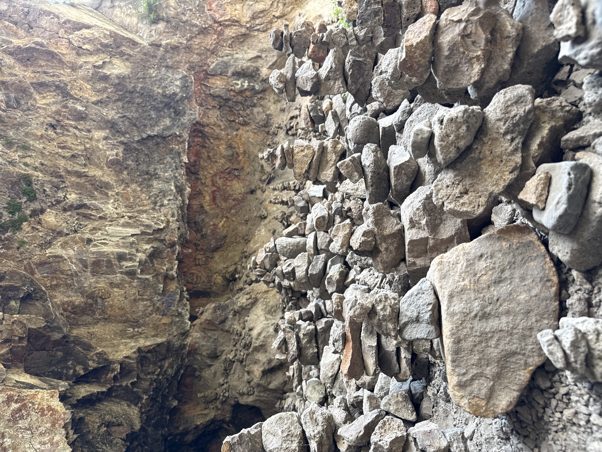 The image depicts a natural rock formation, likely a cave or overhang, in Goght, Armenia. The foreground is dominated by numerous stacked stones, forming cairns of varying heights and densities. These cairns are composed of rough, grey and brown stones of different sizes, with some larger, flatter stones forming bases. In the background, the rock face of the overhang displays a rough, textured surface with natural striations and coloration. Some faint markings, possibly graffiti, are visible on the rock face. The environment appears to be rocky and uneven, with smaller stones and gravel covering the ground. No people or animals are present in the scene.