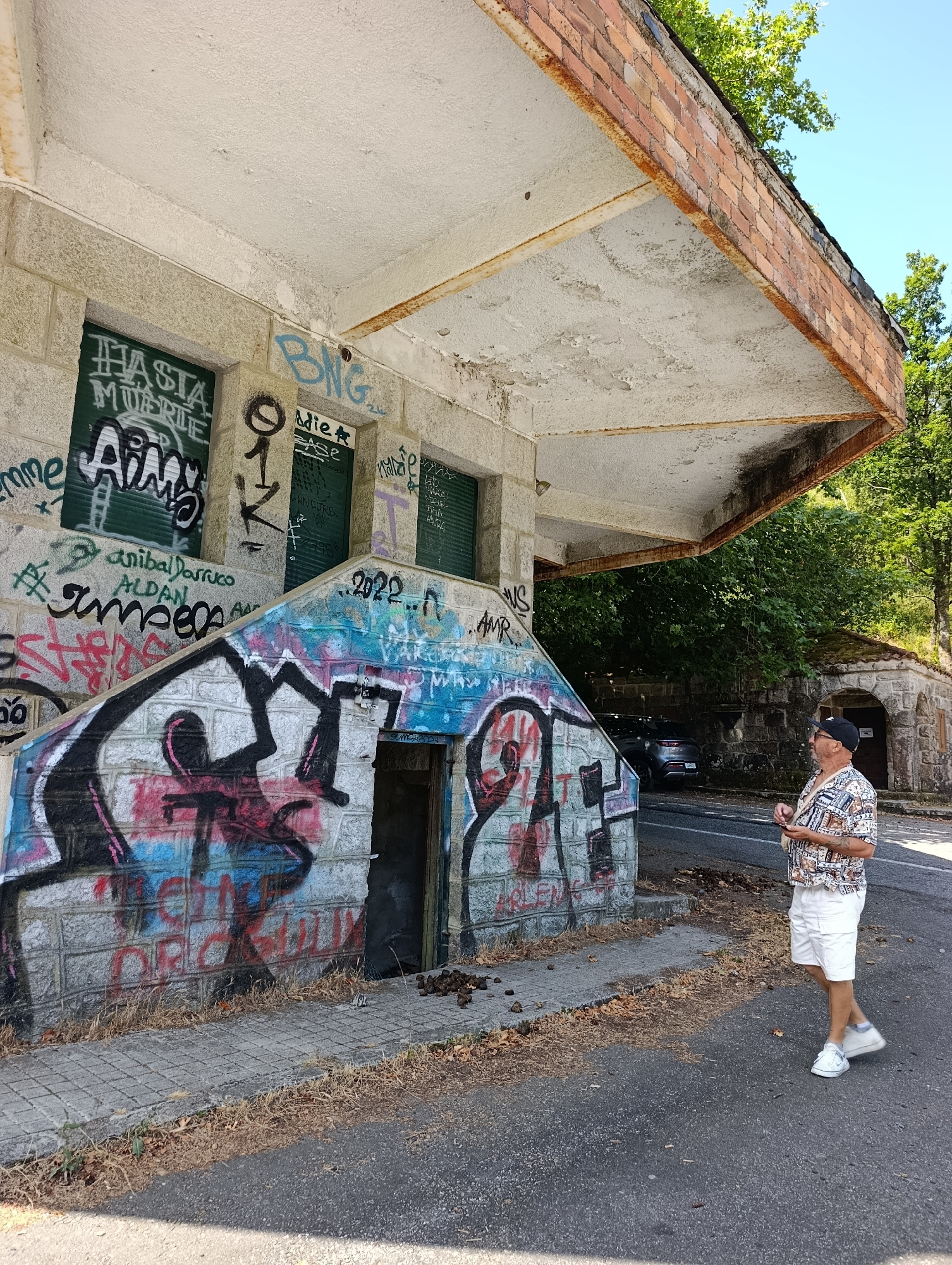 A man stands on a paved area next to a building covered in graffiti. The building appears abandoned, with a decaying roof supported by rusty beams and crumbling concrete. The man is wearing a patterned short-sleeved shirt, white shorts, a baseball cap, and white sneakers. He is looking upwards towards the building's roof, holding a small object in his right hand. The building has a staircase leading up to an entrance, also adorned with colorful graffiti. Beyond the building, trees and a road are visible. The ground is a mix of pavement and dry grass and leaves. The scene appears to be in a rural or semi-urban setting, possibly a derelict structure in Água da Pala, Portugal.