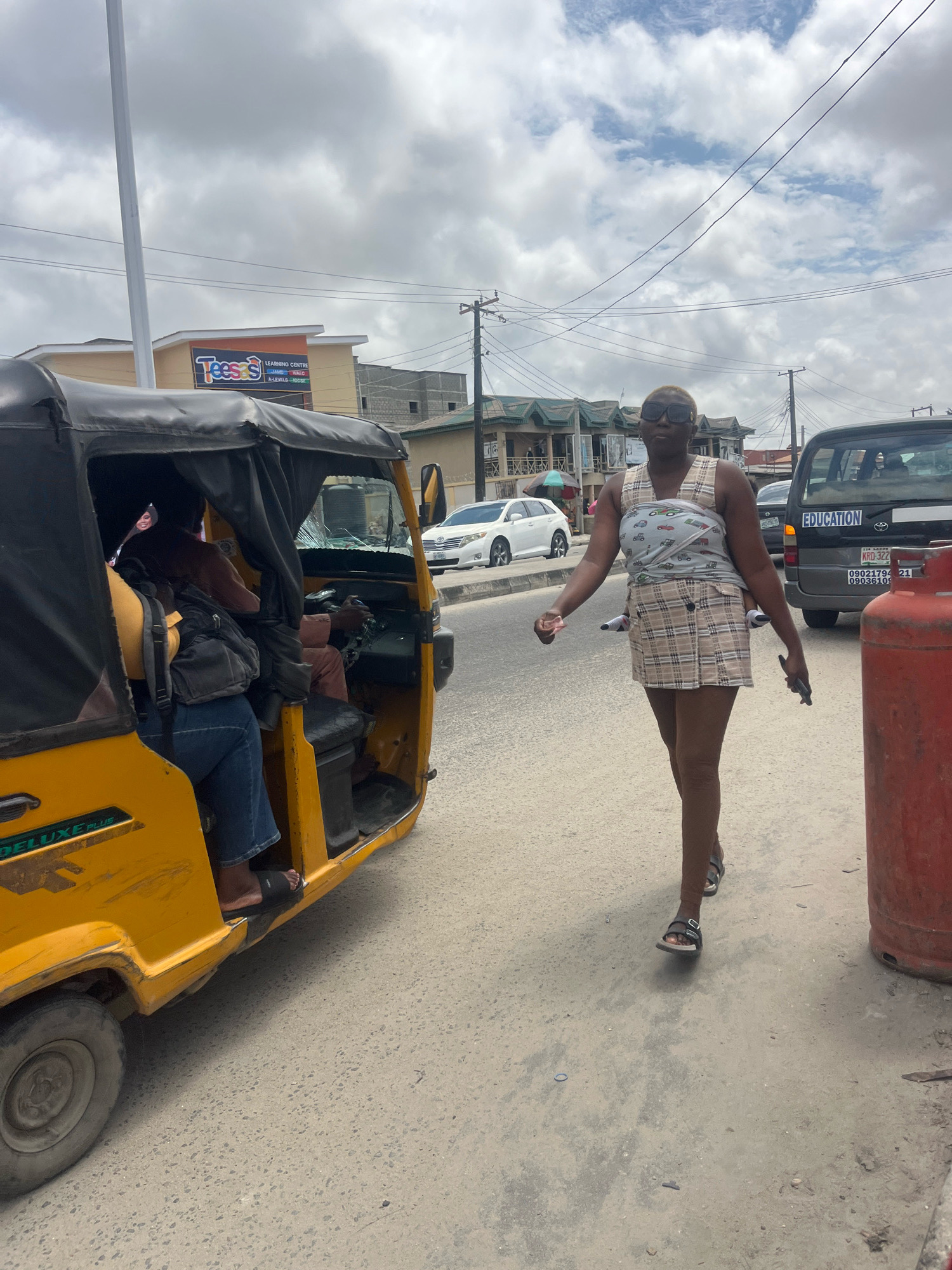 A woman wearing sunglasses, a patterned vest, and a plaid skirt walks on a street in Ado, Nigeria. She carries a dark-colored phone in her right hand. Behind her, a yellow auto-rickshaw is parked, with a person visible inside. A large red gas cylinder stands on the right side of the street. Other vehicles, including a white car and a dark-colored van with "EDUCATION" written on its back, are also present. Buildings line the street in the background. The sky is overcast with clouds.