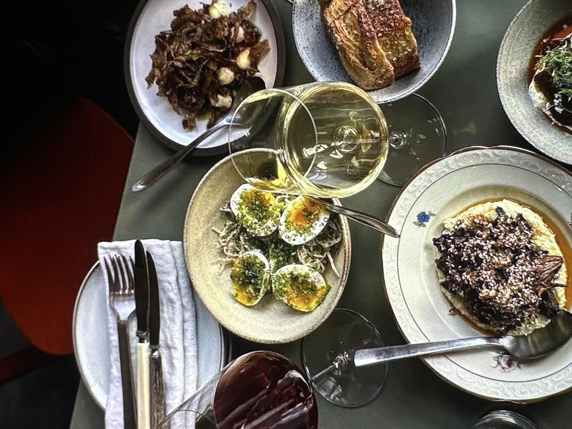 The image depicts an overhead view of a table setting, likely in a restaurant. Multiple plates of food are arranged on a matte green surface.
* One plate contains what appears to be fried onions and garlic.
* Another plate holds slices of bread.
* A third plate showcases sliced boiled eggs with a green herb topping.
* A fourth plate presents a dish with a brown topping on a light-colored base.
* Cutlery is neatly arranged on a white cloth napkin next to an empty plate.
* Two wine glasses are visible: one filled with red wine, the other with white wine and overturned.
The setting suggests a meal shared between multiple individuals. The location (Yerevan, Armenia) cannot be confirmed from the visual information provided.