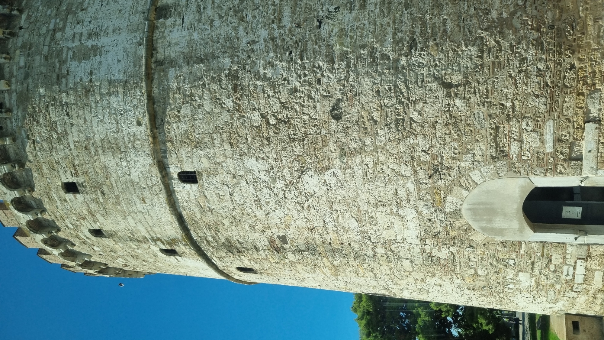 This is a low-angle, close-up shot of the exterior of the White Tower in Thessaloniki, Greece. The tower is constructed of light-colored stone blocks, with a circular base and a crenellated top. Small, rectangular windows are visible at various heights along the tower's wall. An arched entrance with a dark doorway is located at the base. To the left of the tower, a lush green tree and a clear blue sky are visible. The bottom left corner of the image shows a small concrete structure.