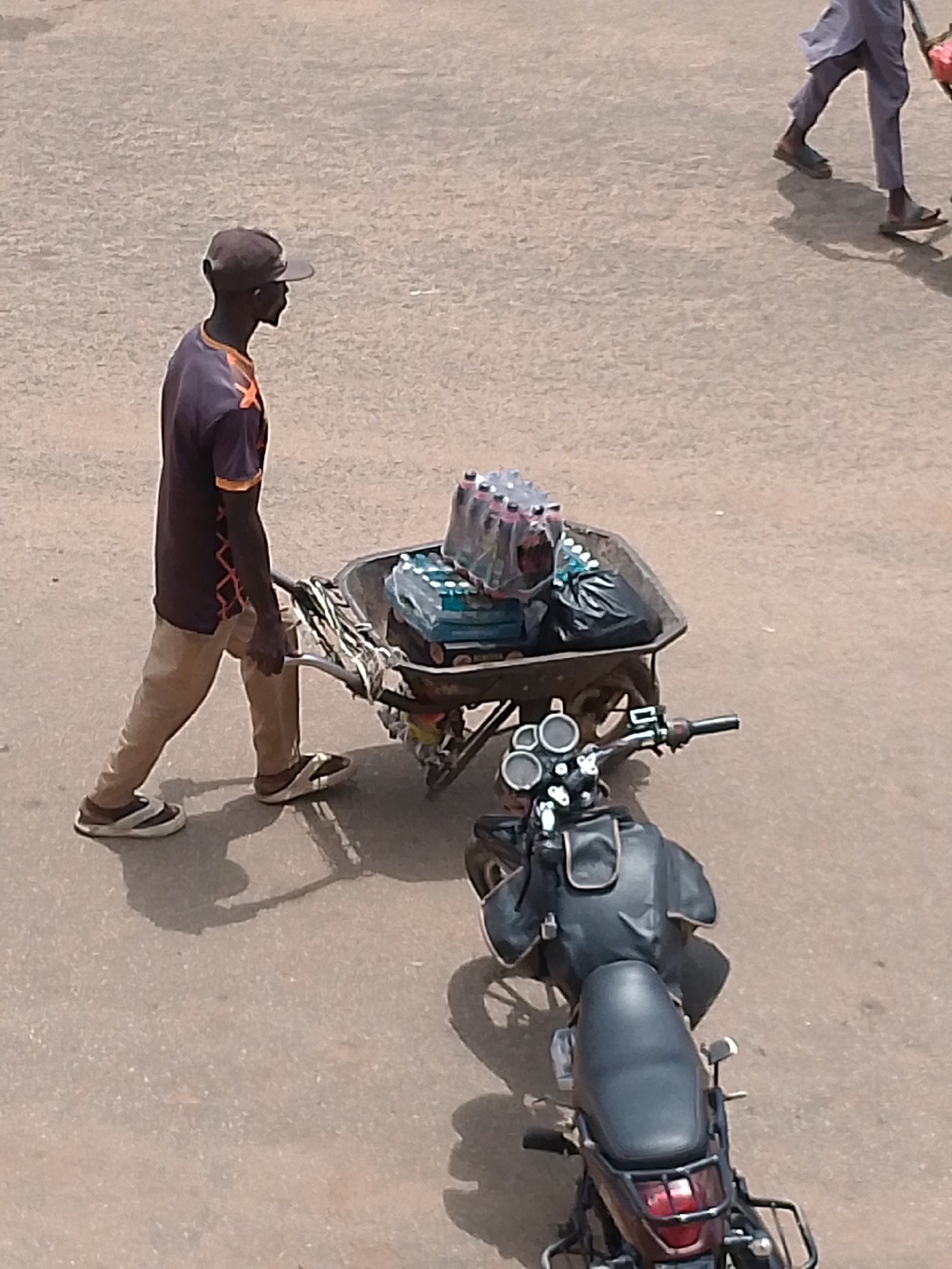 In the city of Bukuru, Nigeria, a man wearing a purple t-shirt with orange accents and a baseball cap pushes a wheelbarrow loaded with bottled drinks and a black trash bag. The wheelbarrow is also attached to a motorcycle, suggesting a method of transport. The ground is paved with asphalt. In the background, another person, partially visible, walks away from the camera, also wearing light-colored clothing. The scene appears to be a street or marketplace.