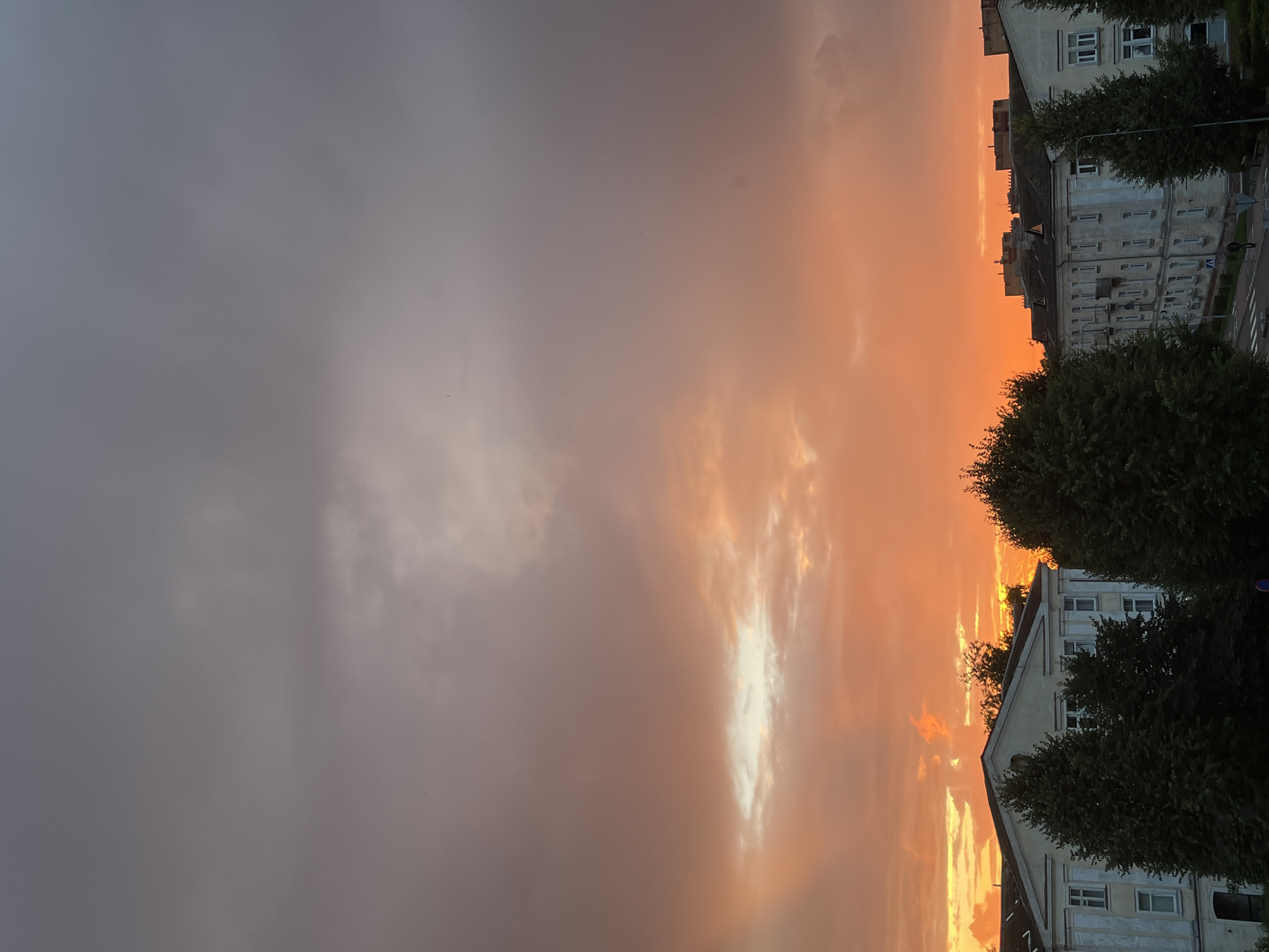 The image depicts a sunset over an urban landscape, likely in Kohtla-Järve linn, Estonia. The sky is filled with dramatic clouds painted in hues of orange, pink, and grey, suggesting a transition from daylight to evening. A bright, illuminated patch in the clouds indicates where the sun is setting or has recently set.
In the foreground, several multi-story buildings with light-colored facades and dark roofs are visible. These appear to be apartment blocks, characterized by numerous windows and balconies. Mature trees, predominantly dark green and dense, are interspersed among the buildings and in front of them, partially obscuring the lower levels of the structures. A street or road is visible in the lower right section of the image, with a crosswalk and some streetlights. One or two figures can be faintly seen walking on the street. The overall impression is of a typical residential area experiencing the end of the day.