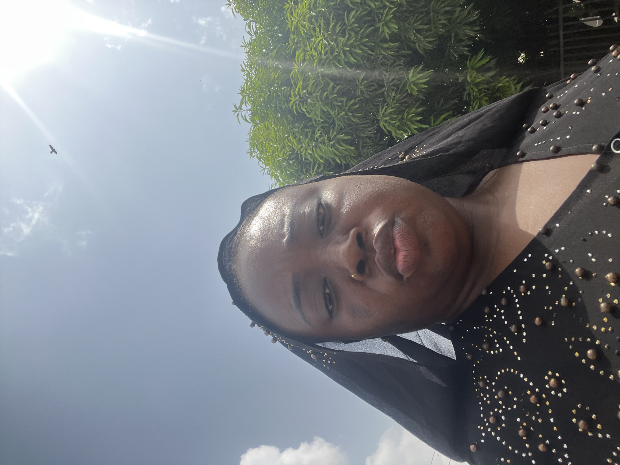 A woman with dark skin, wearing a black top decorated with beads, is looking at the camera with a slightly puckered expression. She has a nose ring on her left nostril. Her head is covered in black fabric. Behind her is a tree with green leaves, a blue sky, and the sun. There are clouds in the sky, and a bird is visible. The photograph was taken outside in daylight. Abuja, Nigeria, is the stated location.