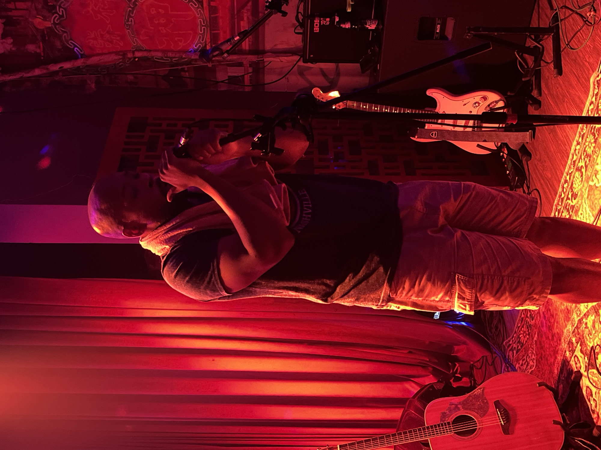 A man stands on a stage in front of a red curtain, speaking into a microphone. He is wearing a dark t-shirt, light-colored shorts, and has a towel draped around his neck. An acoustic guitar rests near the front of the stage, with an electric guitar on a stand behind the man. The room is dimly lit with red lighting. The city of Đà Nẵng, Vietnam cannot be confirmed from the provided image.