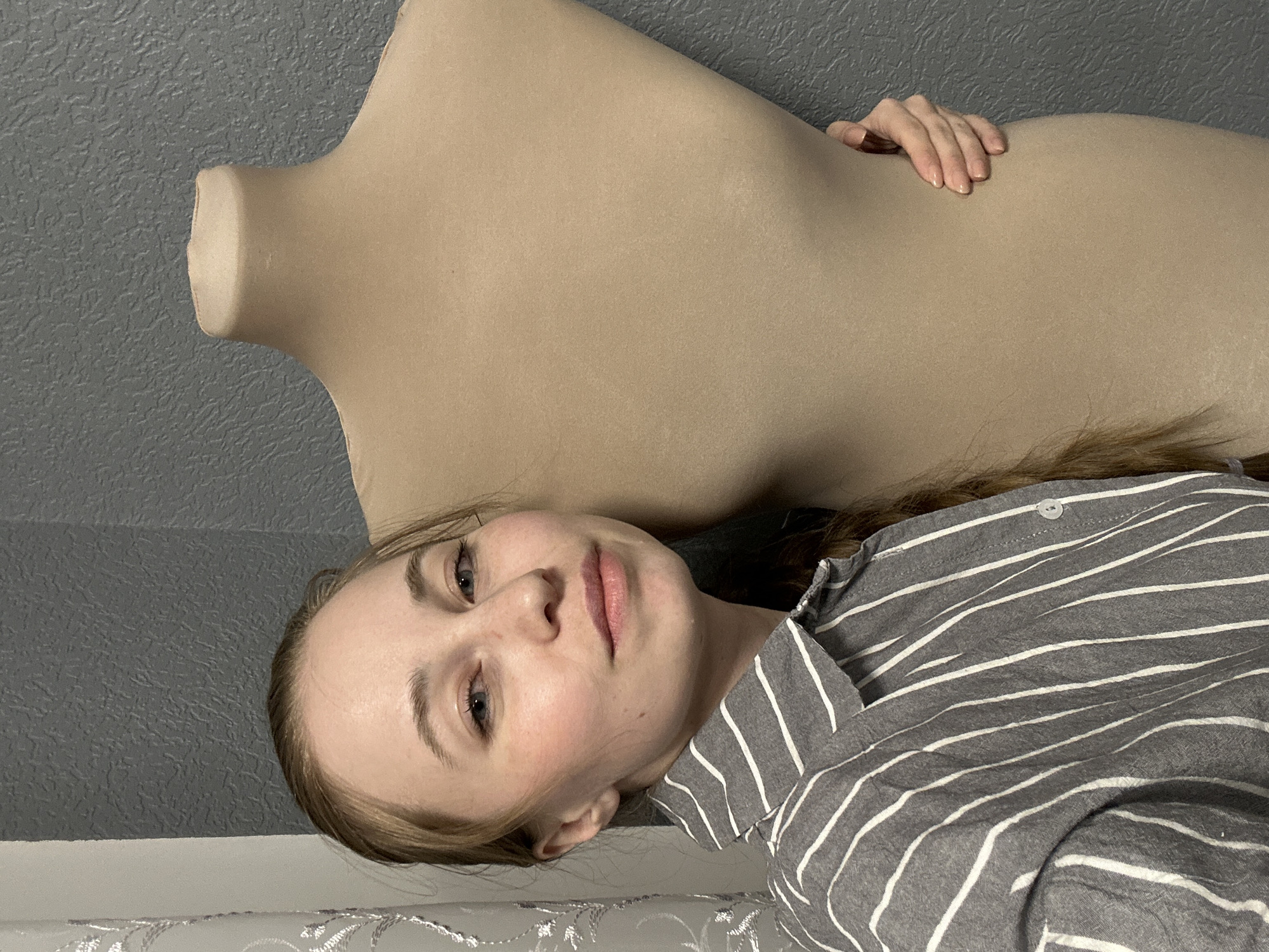 A woman is lying down next to a beige dress form mannequin. The woman has light skin, blue eyes, and light brown hair. She is wearing a gray and white striped shirt. Her hand is resting on the mannequin. The background consists of a gray textured wall. No indication is given that the photo was taken in the city of Saint Petersburg, Russia.
