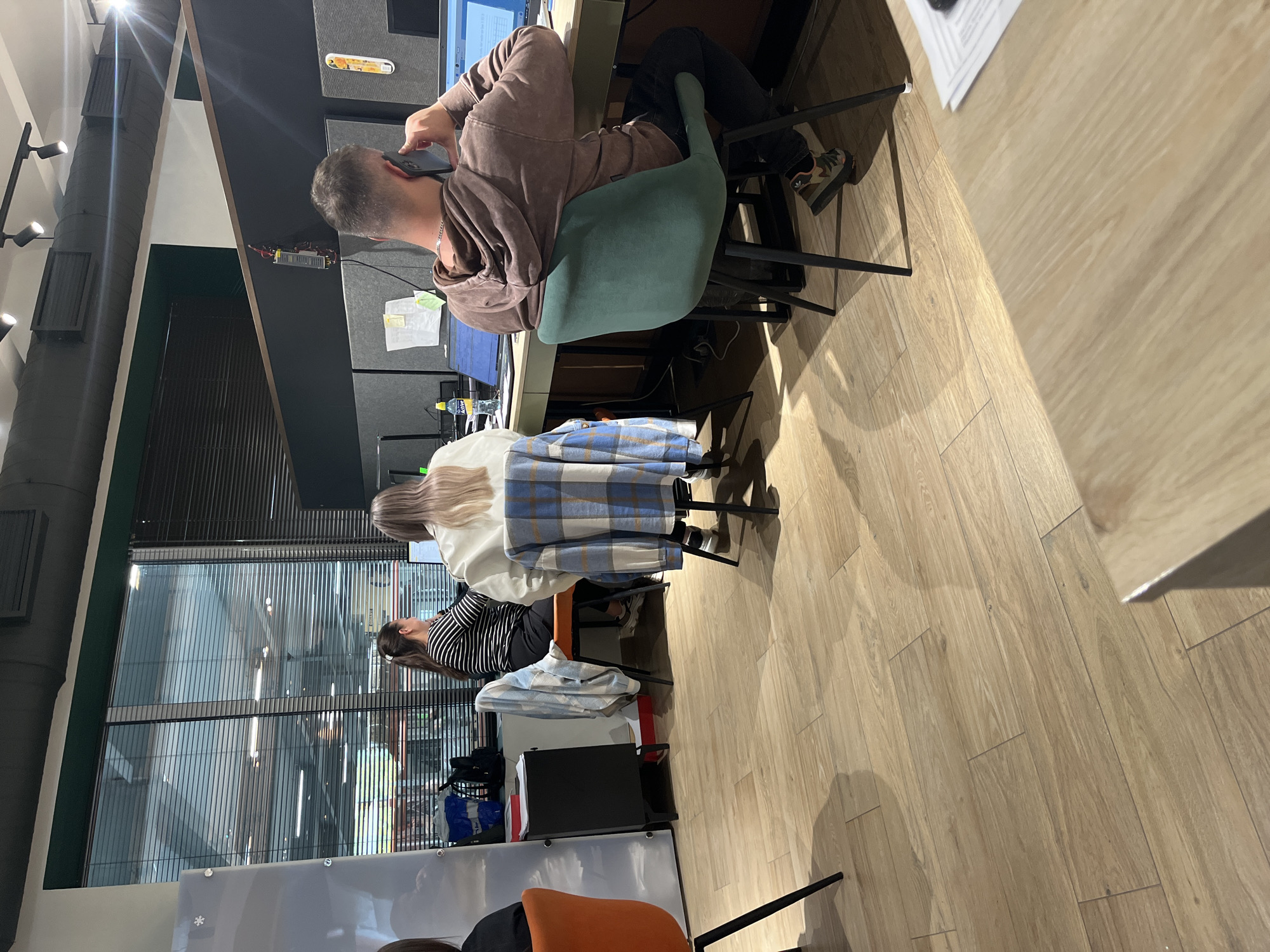 The image depicts an indoor office or coworking space, featuring three individuals seated at their desks. The scene is oriented sideways, but assuming correct orientation, the view is from the back of the room towards the front.
In the foreground, a man with short, dark hair is seen from behind, wearing a brown hoodie. He is seated on a teal-colored office chair and is actively talking on a black mobile phone held to his right ear. His desk is light wood, and a computer monitor, papers, and a small yellow container are visible on its surface.
Further into the room, two women are also seated at their desks, viewed from behind. The woman closer to the foreground has long, straight blonde hair and is wearing a white top; a blue and white plaid shirt is draped over the back of her chair. Behind her, another woman with dark hair is wearing a black and white striped top. A clear water bottle, with the brand name "BORJOMI" partially visible on its label, sits on the desk between the man and the first woman. All individuals appear to be engaged in work or desk-related activities.
The setting includes light wooden flooring and dark grey partition walls or desks. To the left, an industrial-style black ventilation duct runs vertically, featuring several attached spotlights that illuminate the area. A large window with vertical slats or blinds is prominent in the background, showing reflections of interior lighting. The overall impression is that of a modern, functional workspace.
