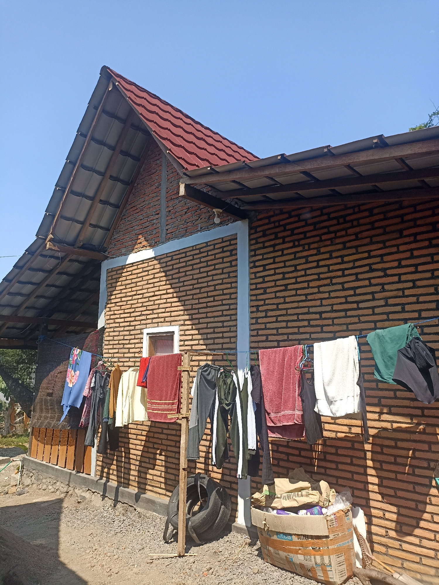 The image depicts the exterior of a brick building under a clear blue sky. The building features a red-tiled gable roof with exposed wooden rafters. Laundry is hung on a clothesline strung across the front of the building, with various garments including shirts, pants, and towels visible. The brickwork of the building is predominantly orange-brown with dark mortar lines, and a light blue band runs horizontally across the upper part of the wall. A small window with a red frame is visible on the wall. In the foreground, the ground is covered with gravel. To the right of the building, a cardboard box filled with various items, including bags and bottles, is placed on the ground. A discarded tire is propped up against a wooden post next to the box. The scene suggests a residential or domestic setting, likely in Mojopanggung, Indonesia, given the architectural style and the bright, sunny weather. There are no people visible in the image.