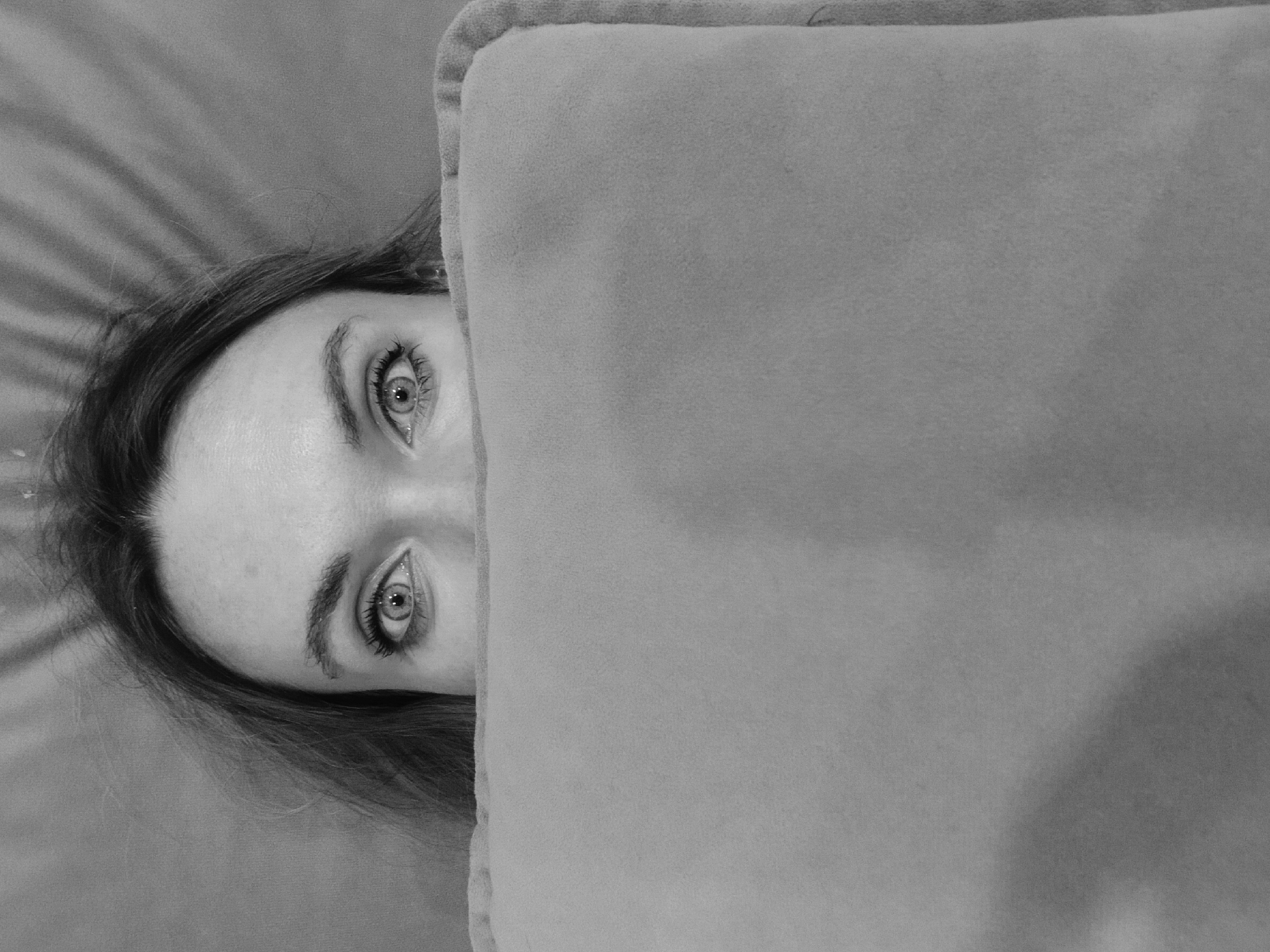 A black and white close-up shot shows a person partially obscured by a rectangular object. The person's face is visible, including their eyes, forehead, and part of their hair. The rectangular object, likely a pillow or blanket, is positioned to the right of their face, covering the majority of their body. The background appears to be a plain, slightly textured surface. There is no indication of the location being Votkinsk, Russia in the image.