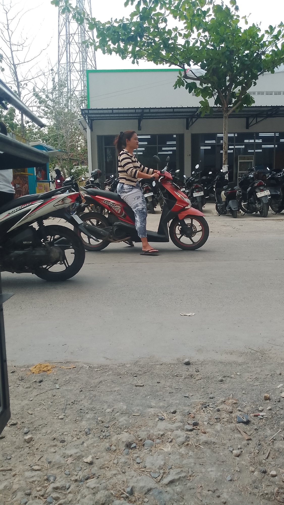 A woman with dark hair pulled back in a ponytail is sitting on a red Honda Beat scooter. She is wearing a striped long-sleeved shirt and patterned leggings, with flip-flops on her feet. The scooter is parked on a paved road in front of a building with a green and white facade, featuring large glass windows and a dark awning. Behind the building, a tall metal tower and trees are visible. A white and red scooter is parked to the left of the red scooter, and several other parked scooters are visible in the background. The ground in the foreground is a mix of gravel and dirt, with some scattered debris. The scene appears to be in the city of Karanglangu, Indonesia.