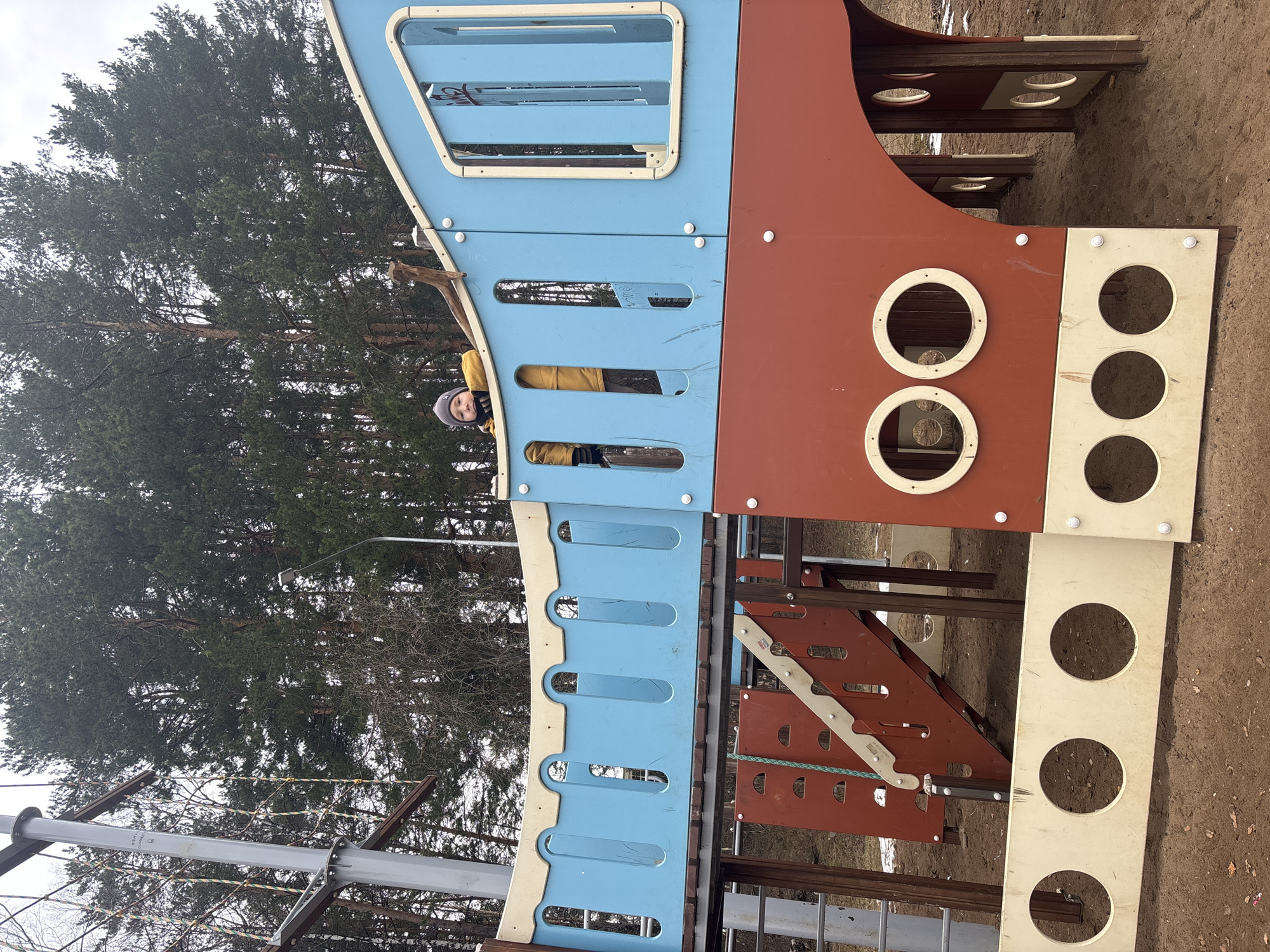 A young child wearing a yellow jacket and grey hat looks out from the blue section of a playground structure. The structure has a combination of blue and reddish-brown paneling with white circular cutouts. Trees are visible in the background. The location is in the city of Chaikovsky, Russia.