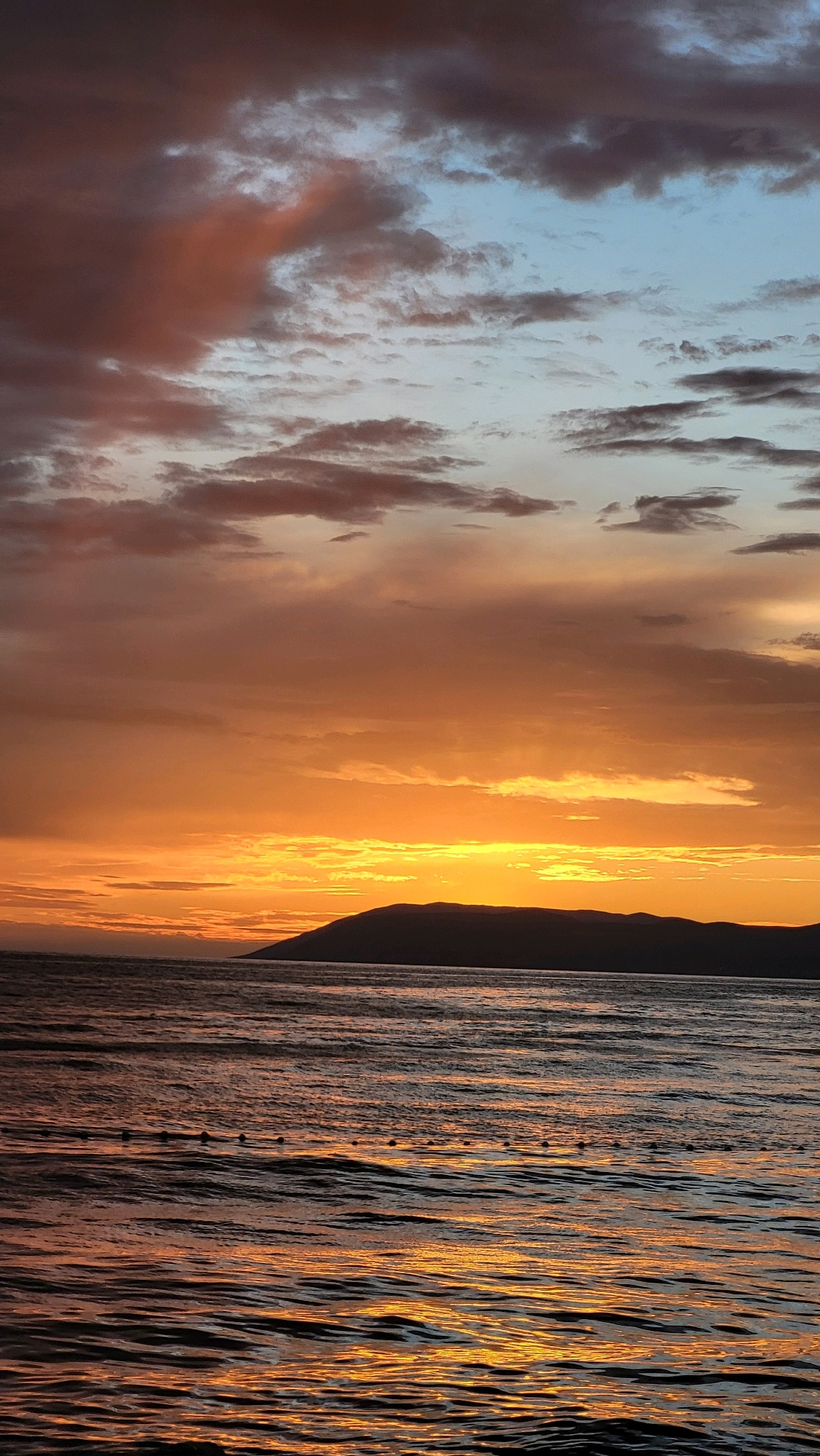 This image depicts a sunset over the Adriatic Sea, likely taken from the coastline of Općina Tučepi, Croatia. The sky is a dramatic canvas of colors, with hues of deep orange, yellow, and pink dominating the horizon, transitioning to lighter blues and grays towards the top. Wispy clouds are scattered across the sky, reflecting the warm tones of the setting sun. A dark, silhouetted mountain range forms the backdrop, stretching across the horizon. The sea in the foreground is choppy, with ripples reflecting the vibrant colors of the sunset, creating streaks of orange and gold on the dark water. In the mid-ground of the water, a series of small, dark buoys are visible, arranged in a line, possibly marking a swimming area or a fishing net. The overall atmosphere is serene and visually striking, capturing the beauty of a coastal sunset.