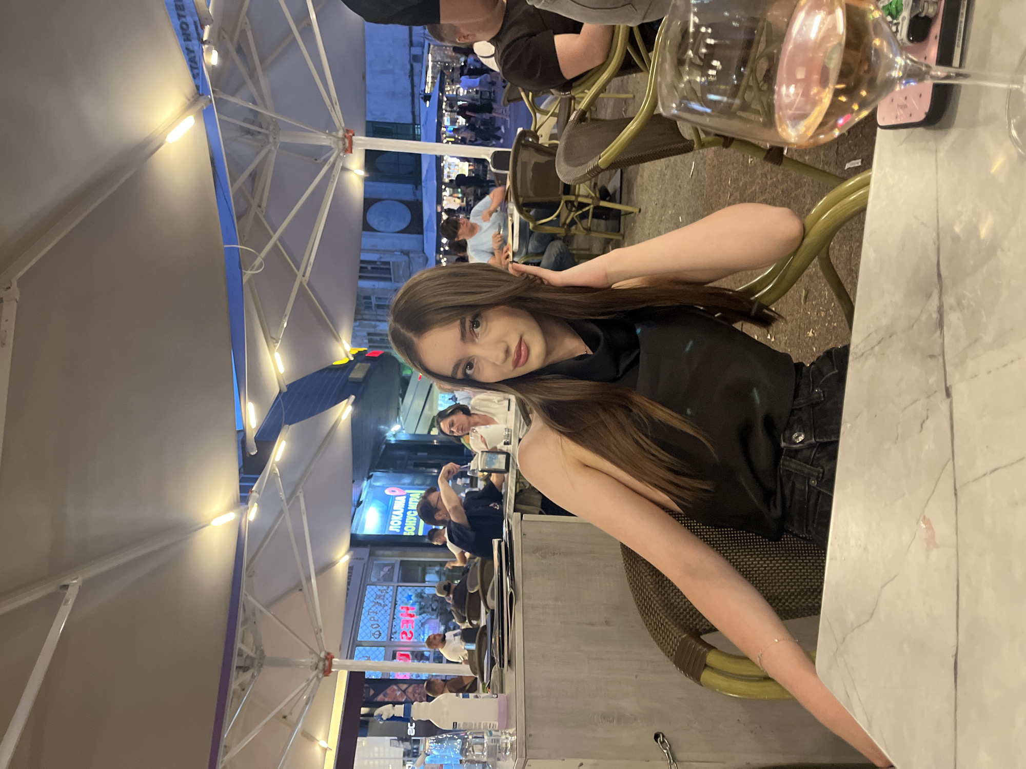 A young woman with long brown hair sits at an outdoor cafe. She wears a black top and dark jeans. A glass of wine is on the table next to her. People are seated at other tables under large, illuminated umbrellas. Shop signs in Cyrillic script are visible in the background, indicating a location in Bulgaria, specifically Plovdiv.