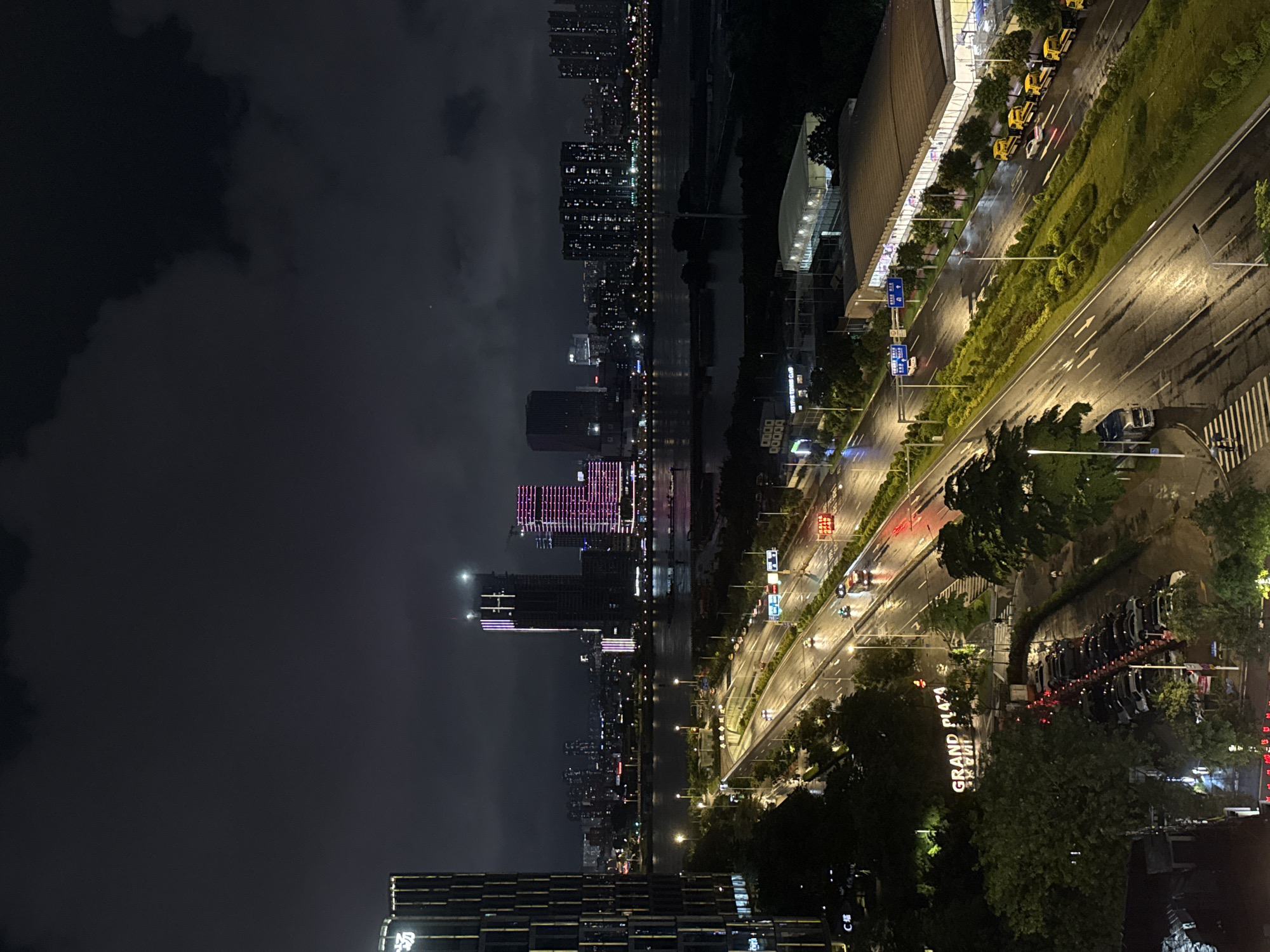This nighttime aerial view depicts a section of Haizhu District, Guangzhou, China, after rain. The scene is dominated by a multi-lane, divided highway running from the lower left to the upper right, its wet surface reflecting the urban lights. Numerous vehicles, including cars, trucks, and a line of yellow utility vehicles, are present on the road and in a parking lot. Several pedestrians are visible on sidewalks and a crosswalk, one pair beneath an umbrella.
On the left side of the foreground, a tall, modern building with a distinct patterned facade stands prominently, displaying the illuminated "GRAND PLAZA" sign. Below it, a densely packed parking lot holds rows of parked cars. Adjacent to the road on the right, a large structure with a curved, brown roof and a sign for "OLYMPIC TENNIS CLUB" is partially visible. The median and road edges are lined with trees and manicured shrubbery.
Beyond the highway and green spaces, a wide body of water, likely the Pearl River, reflects the lights from the distant cityscape. Across the river, a dense array of high-rise buildings forms the skyline. Notable among these are two distinct structures: one under construction, illuminated with vertical white and purple lights, and another to its right, displaying prominent pink and purple horizontal lighting patterns. The sky above is dark and overcast with visible cloud formations.