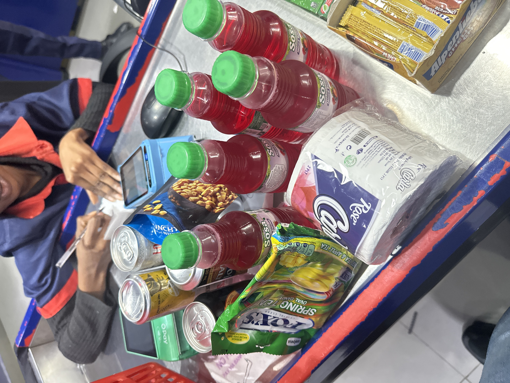 A cashier in Barden Yana, Nigeria, is processing items at a checkout counter. The cashier, wearing a blue uniform with red trim, is writing on a piece of paper. On the counter, various products are laid out, including several bottles of red-colored beverages with green caps, cans of drinks, a packet of "Sosa" fruit drink, a pack of "Rose" tissues, and a box of candies labeled "Alpenliebe." A green payment terminal is also visible. The counter surface is metallic and shows signs of wear.