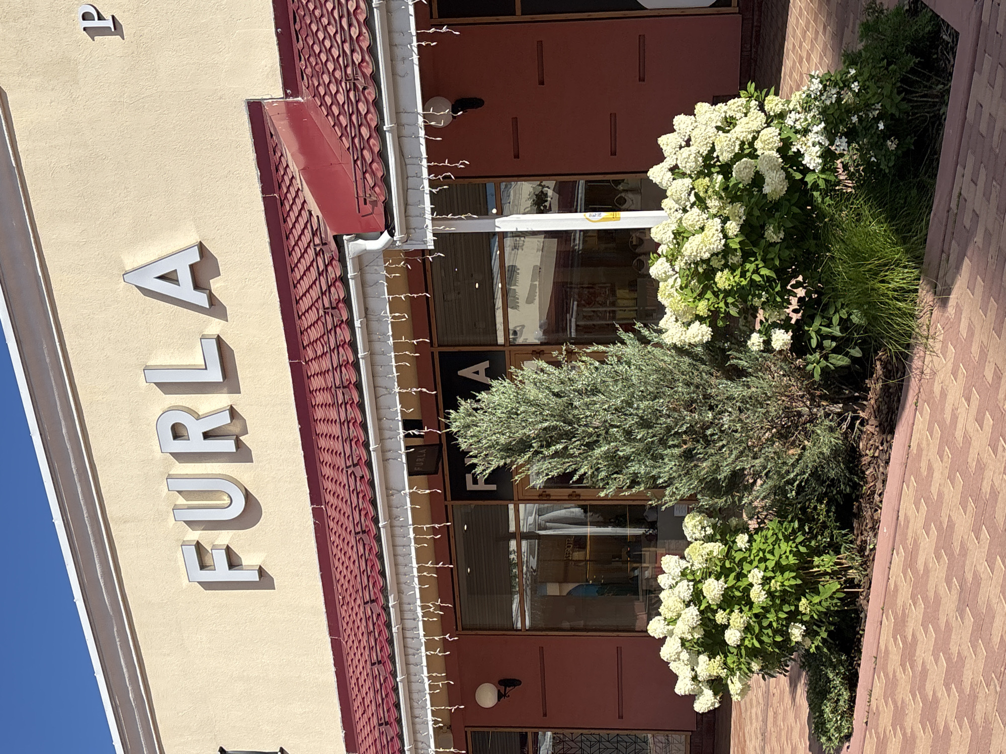 A storefront with the name "FURLA" displayed prominently in large, white, three-dimensional letters on a beige facade. Below the name, a red tiled roof with a white gutter system is visible, adorned with decorative white string lights. The entrance to the store features large glass windows and doors. In front of the store, a conifer tree stands beside a large bush with white flowers, and a smaller flowering bush with white and green foliage. The ground in front of the storefront is paved with red brickwork. A single white globe light fixture is mounted on the exterior wall to the left of the entrance, and another to the right. A sign with "FURLA" in white letters on a black background is visible above the entrance. A partial "P" is visible on the far right of the building. The sky is clear and blue. The scene depicts the exterior of a retail establishment, likely in a shopping area, with landscaping elements.