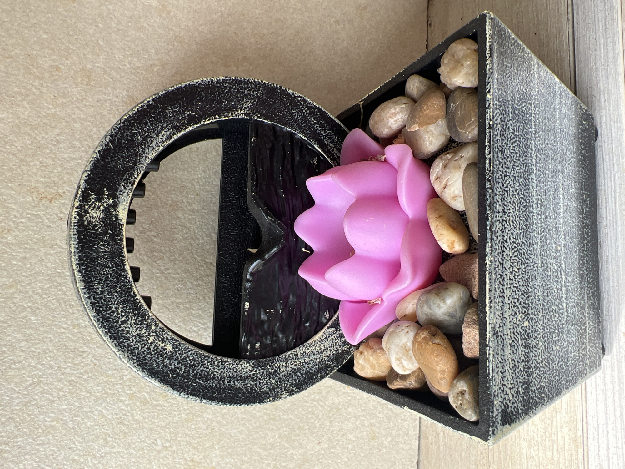A small, self-contained decorative water feature is centrally positioned on a light-colored wooden surface. The object comprises a dark grey, tapered rectangular base with a mottled, textured finish that includes flecks of lighter grey or white. This base is filled with an assortment of smooth, rounded pebbles in natural hues of beige, brown, and various shades of grey. Resting amidst the pebbles is a prominent, bright pink, lotus-shaped object, likely a candle or a static decorative element. Behind the lotus, a dark grey, arch-shaped structure rises, matching the mottled, textured finish of the base. The interior surface of this arch presents a smooth, dark, undulating texture, suggestive of a small water reservoir or a miniature waterfall. Several small, black, cylindrical protrusions are visible along the lower edge of the arch. The background consists of a uniformly textured wall in a light beige tone. No individuals or specific actions are depicted in the scene. The precise location within the city of Burgas, Bulgaria, cannot be determined from the visual information provided.