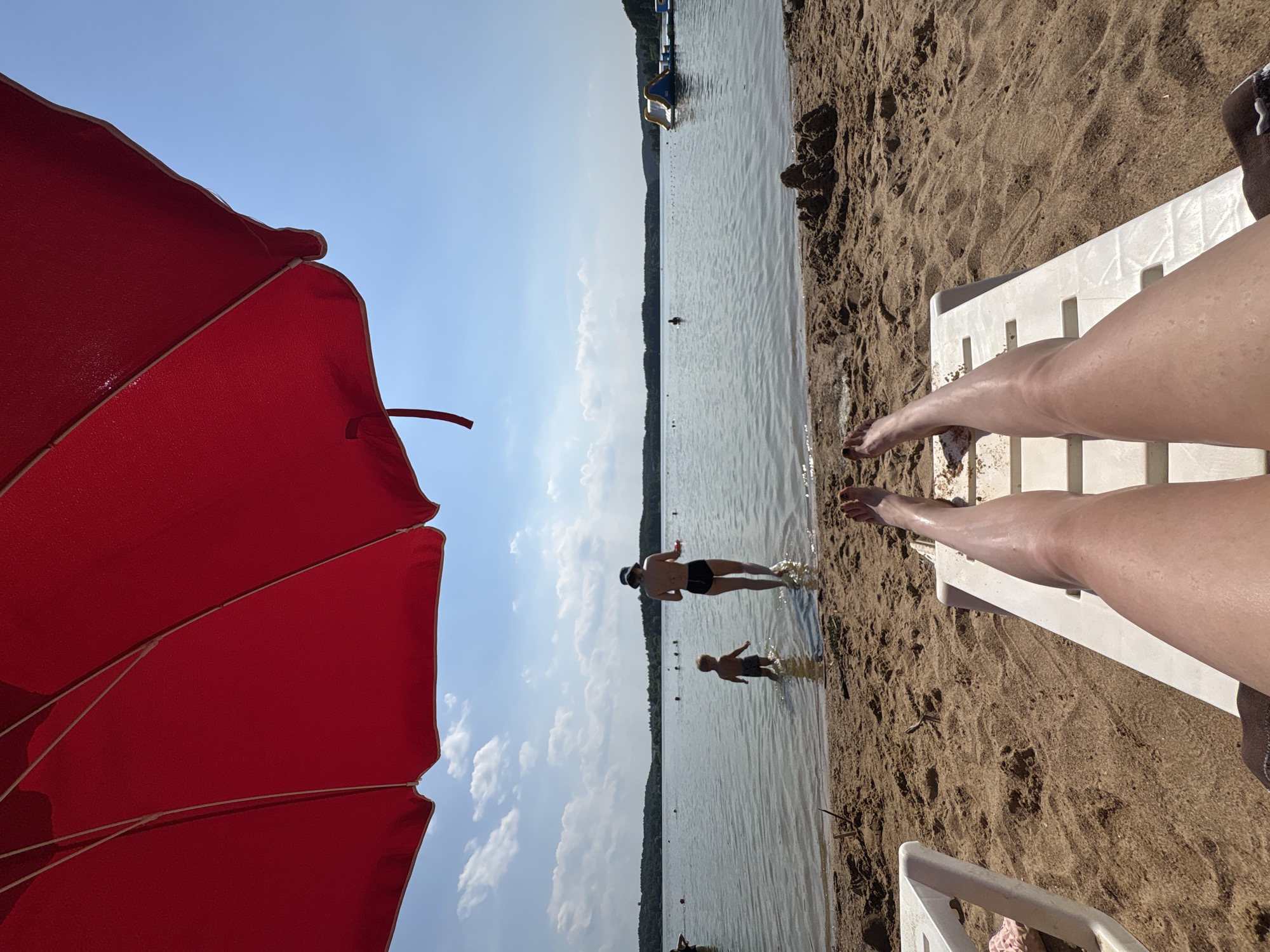 A person's legs are stretched out on a white lounge chair on a sandy beach under a large red umbrella. The beach is adjacent to a body of water, likely a lake, where other people are visible. In the water, a child stands near an adult who is facing away from the camera. Further out, there are several other people swimming and an inflatable water structure. The background shows a tree-lined horizon under a bright blue sky with scattered clouds. The scene appears to be a recreation area in the city of Ozyaritska-Slabadski rural council, Belarus.