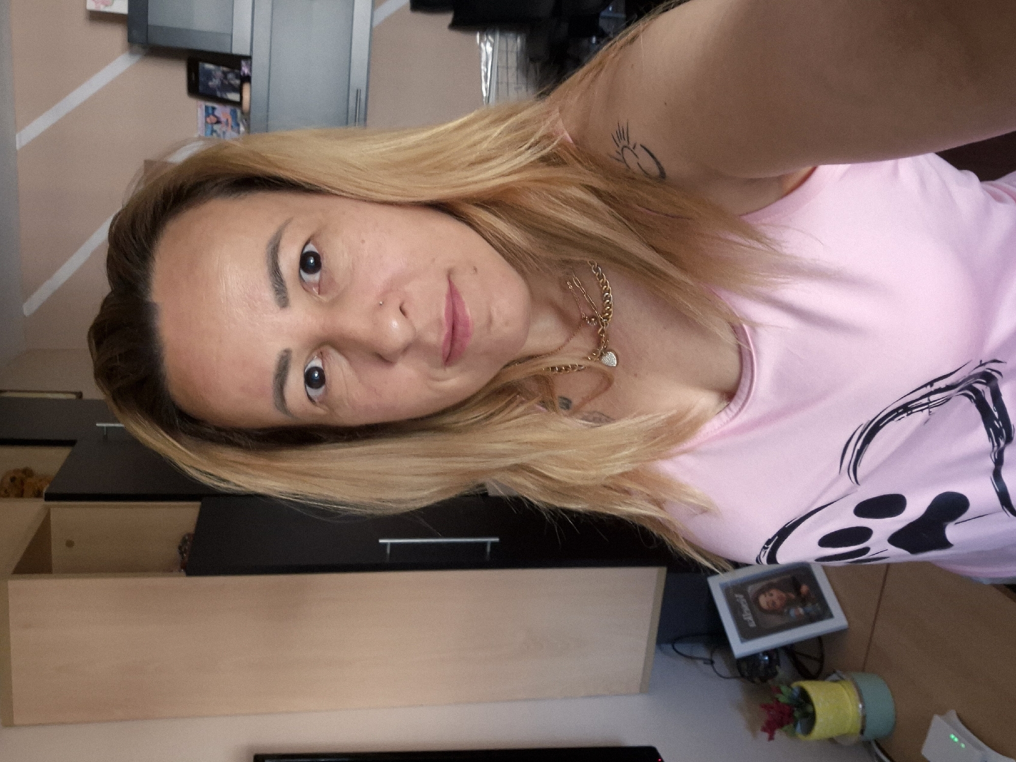 A medium shot captures a woman with blonde hair, wearing a pink tank top with a black paw print design, as she stands in an indoor setting that appears to be a home office. The woman is positioned slightly to the right of the frame, facing the camera directly. Her expression is neutral. Her left arm is visible, with a tattoo of a sun symbol on her shoulder.
In the background, to the left, is a tall, two-toned cabinet with a light wood grain on the left side and a dark brown or black door on the right. To the right of the woman, a desk is visible with a small potted plant with red flowers in a yellow container and a framed picture of a young girl. Further to the right, a dark gray cabinet is visible, with several framed photos on top. The wall behind the desk and cabinets features a diagonal striped pattern in white and a pale pink or beige color. The overall environment suggests a domestic setting. The lighting appears to be natural, coming from an unseen window.