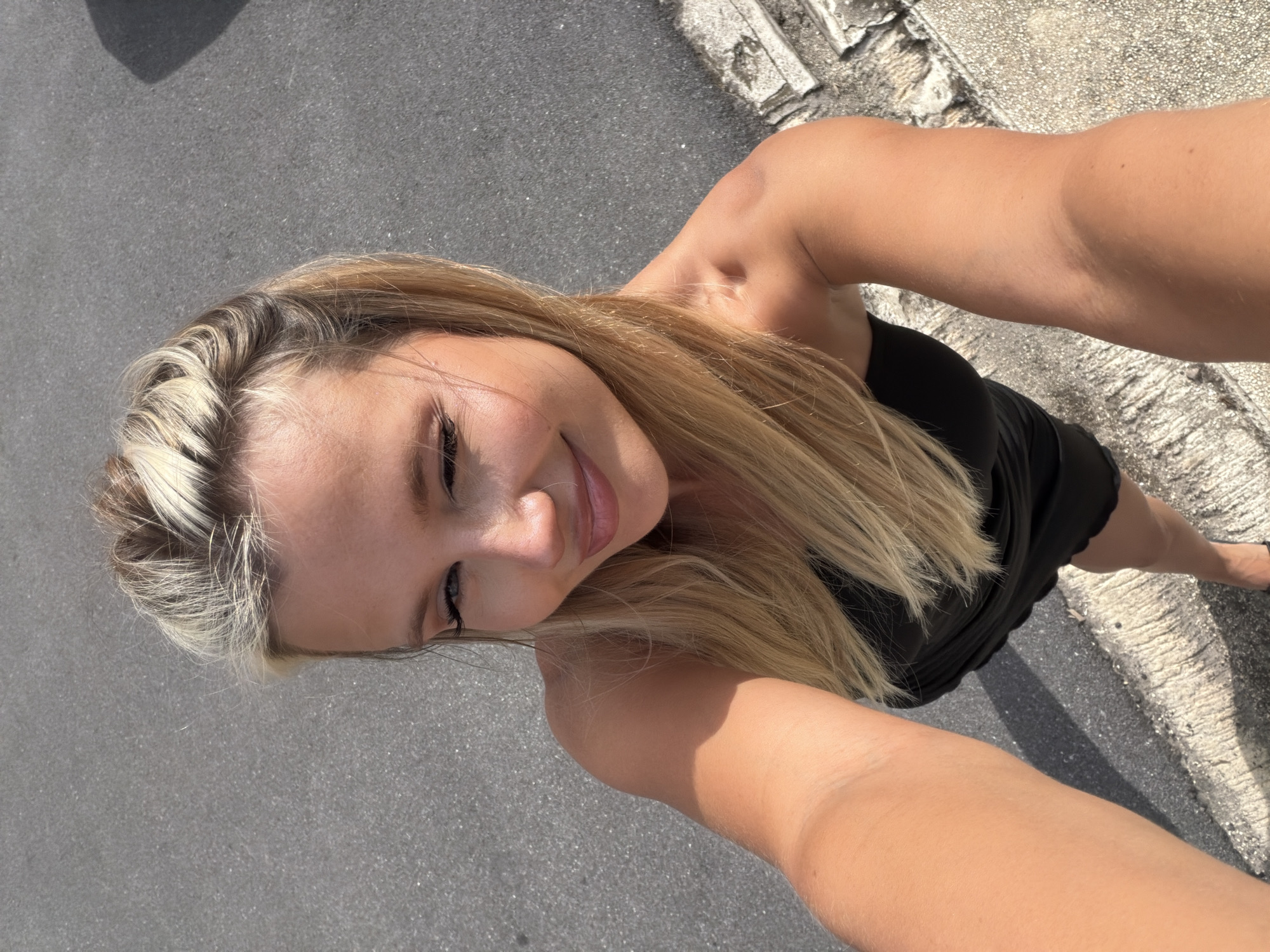 This is a selfie of a young woman with blonde hair and light skin. She is wearing a black dress and is standing outdoors. The sun is shining, casting shadows on the ground, suggesting it is daytime. The background appears to be a paved surface next to a textured, concrete structure. Her facial expression is relaxed and pleasant, with a slight smile. The image appears to be taken on a sunny day, possibly in a warm climate given the subject's attire and the overall lighting.