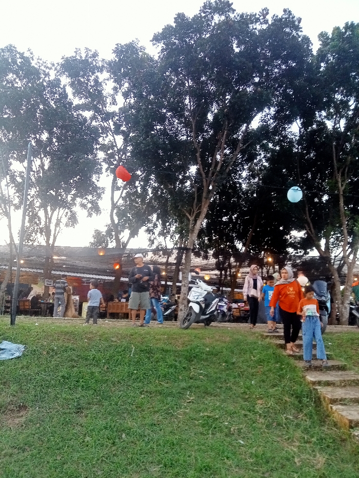 The image shows a group of people standing in front of a restaurant in Jakenan, Indonesia, at latitude -6.758 and longitude 111.075. The restaurant has a wooden roof and is surrounded by lush green grass. The people are all dressed in casual clothing, and some are holding onto balloons. There are a few motorbikes parked near the restaurant. The sky is overcast, and the trees are providing shade. The people appear to be in a relaxed mood. Some people are walking toward the camera while others are standing and talking. Two blue balloons are floating in the trees behind the people. There is a red balloon floating in the trees to the left. There is an unidentifiable object on the grass in the foreground.