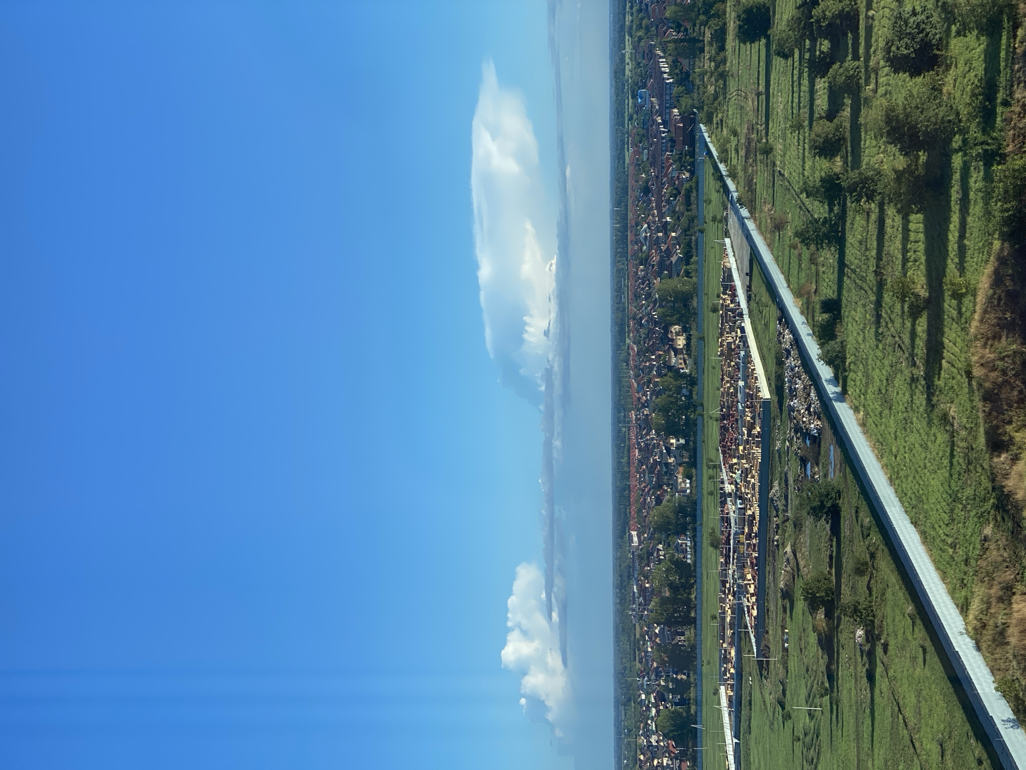 This aerial photograph, taken from a high vantage point, captures a wide expanse of a developing urban or suburban area under a bright, clear blue sky with scattered white clouds. The scene is divided into distinct zones. The uppermost portion of the image is dominated by the sky, featuring a lenticular cloud formation that is prominent and elongated.
Below the sky, a vast residential area unfolds. It consists of numerous houses with red-tiled roofs, densely packed together, creating a uniform pattern across the landscape. Green trees are interspersed throughout this residential zone.
In the middle ground, a construction site is visible. It appears to be a large area with foundations laid for new buildings and stacks of construction materials, primarily wood. This site is demarcated by fences. Adjacent to this construction area, and visible in the foreground, is a long, light-colored barrier or wall that stretches diagonally across the frame. This barrier separates a neatly manicured grassy area from a more rugged, unkempt field with dry vegetation.
The lower foreground consists of green fields with scattered trees and some areas of disturbed earth, possibly indicating ongoing landscaping or construction activity. A large pile of debris, possibly construction waste, is visible along the base of the barrier in the middle ground. The lighting suggests it is daytime, likely morning or late afternoon, given the long shadows cast by the trees. There are no visible people or vehicles in this scene. The overall impression is one of development and expansion within a relatively flat geographical area.
