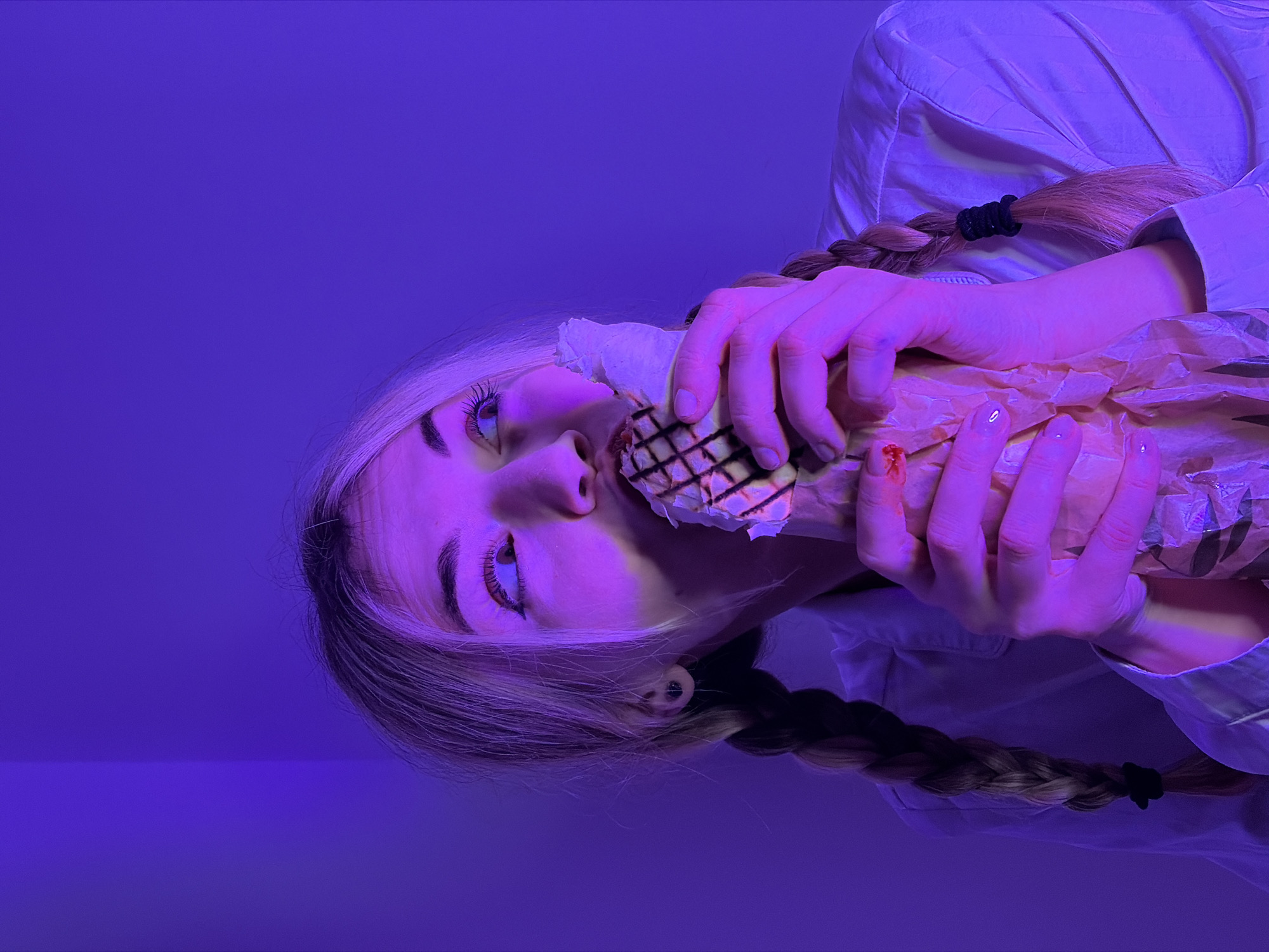The image displays a close-up, rotated portrait of a person, likely female, engaged in eating, set against a vibrant, uniform purple-blue background. The entire scene is bathed in a strong purple-blue light, giving everything a distinct cool and stylized hue.
The person has long, blonde hair, styled into two distinct braids, one of which is clearly visible extending towards the bottom of the frame, secured with a black hair tie. Their skin appears fair but is heavily tinted by the purple lighting. One eye is prominent, featuring a blue or purple iris, and is looking slightly upwards and to the left. Parts of their nose and mouth are also visible, with their mouth slightly open around a food item, suggesting they are in the process of biting or eating.
The food item appears to be a wrap or a sandwich, partially enclosed in a light brown paper wrapper. The visible part of the food has a distinct grilled or waffle-like pattern on its surface. Both hands are holding the food item, bringing it close to their face. A notable detail is a small red mark, resembling blood, on one of the fingers (possibly the index or middle finger) of the hand holding the wrapper.
The person is wearing a light-colored, long-sleeved shirt, possibly white, which also takes on the pervasive purple tint from the lighting. The background is a plain, solid purple-blue, devoid of any discernible features or textures, indicating either a studio setting or a room entirely lit with colored lights. There is no visible text present in the image. The overall impression is one of an intimate, stylized shot, focusing on the act of consumption under dramatic lighting.