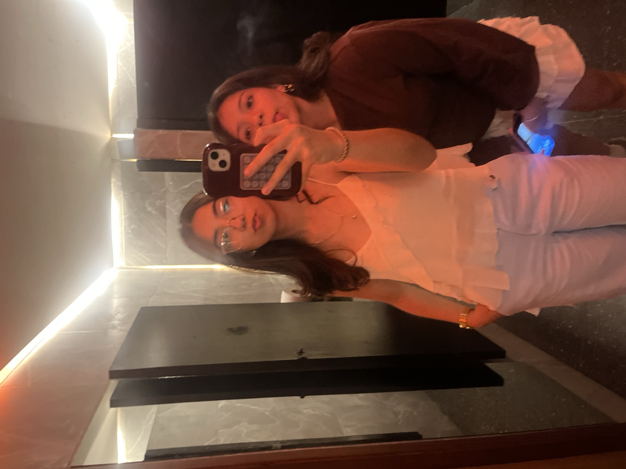 Two young women are taking a mirror selfie in a bathroom. One woman, reflected in the mirror, wears a white top and light-colored pants, holding the phone with a burgundy case. The other woman, also reflected, wears a brown top and white shorts. A cell phone with a blue screen is visible in her hand. The bathroom has a modern design with marble-like tiles, a dark countertop, and recessed lighting along the wall and underneath the countertop. Based on metadata, this image was taken in the city of Puebla, Mexico.