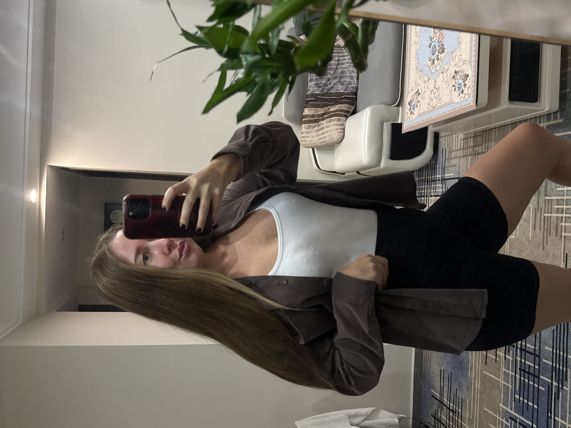 A young woman with long, brown hair is taking a mirror selfie in a room that appears to be in Yiwu, China. She is wearing a white tank top, a brown unbuttoned shirt, and black shorts. She is holding a dark red smartphone up to take the picture. In the background, a grey couch with decorative pillows is visible, as well as a patterned rug on the floor. A plant is in the foreground, partially obscuring the reflection of the room.