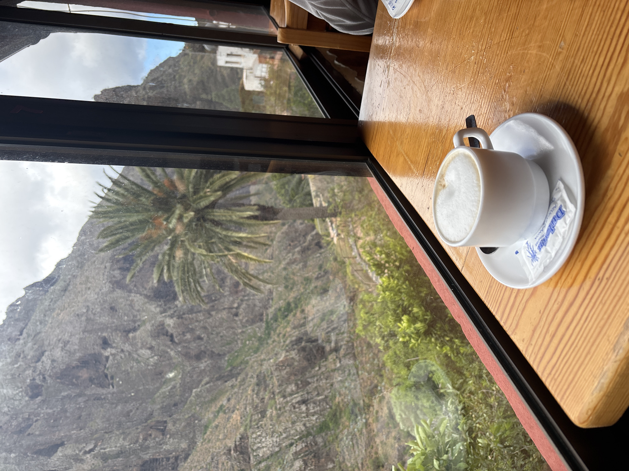 The image depicts a cup of coffee on a saucer with a sugar packet placed on a light brown wooden table, viewed from an elevated perspective. The setting is indoors, possibly a café, with a window providing a view of the mountainous landscape. A large palm tree stands prominently in the middle ground. In the distance, there are houses that may be located in the city of Buenavista del Norte, Spain. The window frame is visible in the foreground.