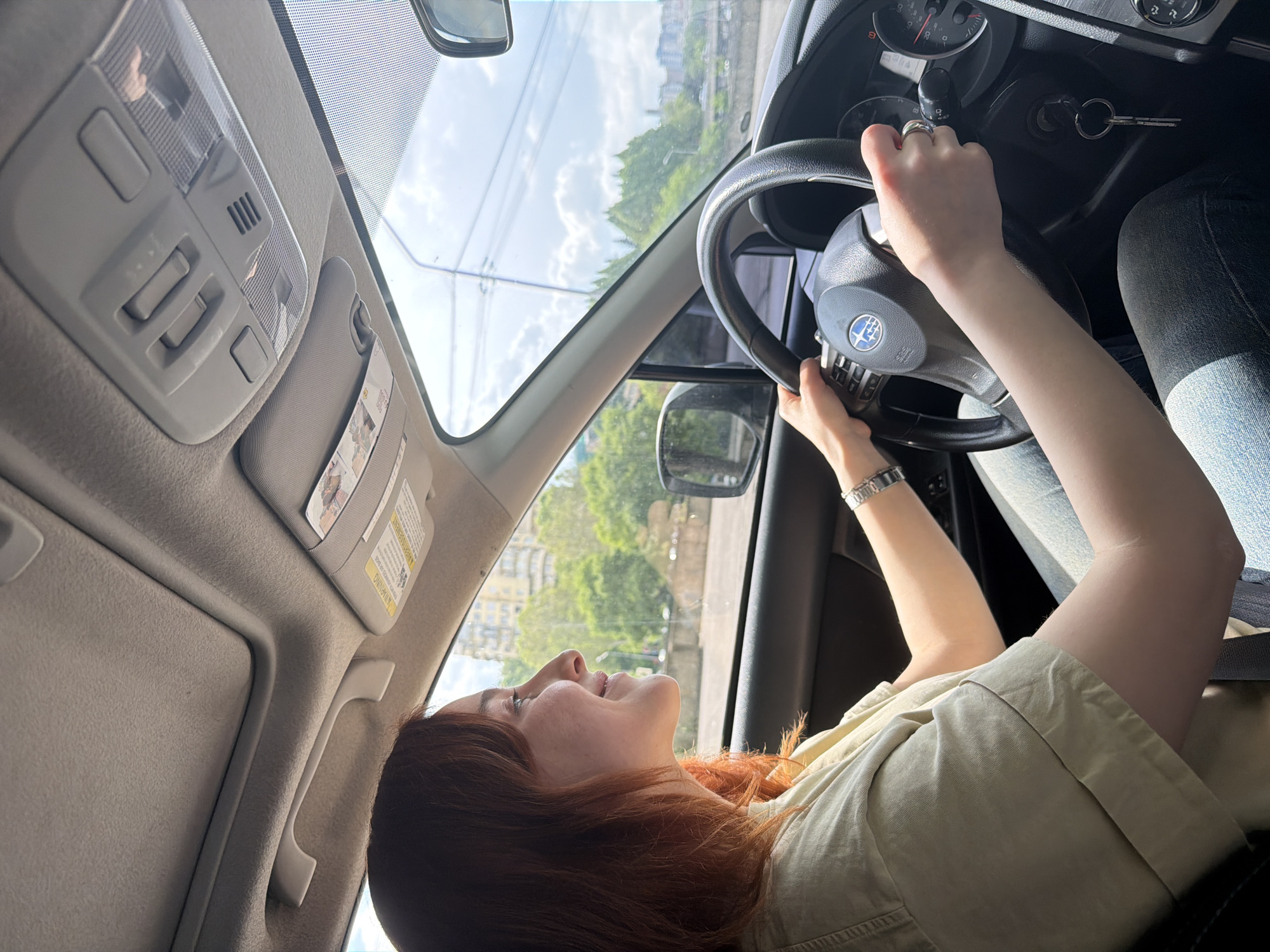 A woman with reddish hair is sitting in the driver's seat of a car, looking up and smiling. She is holding the steering wheel with both hands. Her left hand has a silver ring, and a silver wristwatch is visible on her left wrist. The car is a Subaru, based on the emblem on the steering wheel. The key is in the ignition. Outside the car window, buildings and trees are visible. The scene appears to be in Tbilisi, Georgia, based on the architectural style of the visible buildings and the presence of lush greenery.