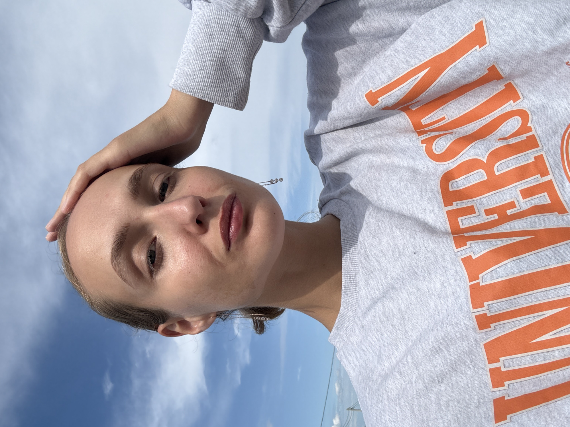 A young woman with blonde hair pulled back is taking a selfie outdoors. She is wearing a light gray sweatshirt with "UNIVERSITY" printed in orange across the chest. Her right hand is touching her forehead, and she is looking directly at the camera with a slight pout. The background is a bright blue sky with scattered white clouds. No specific location in the city of Подшивалово, Russia, can be determined from the image.