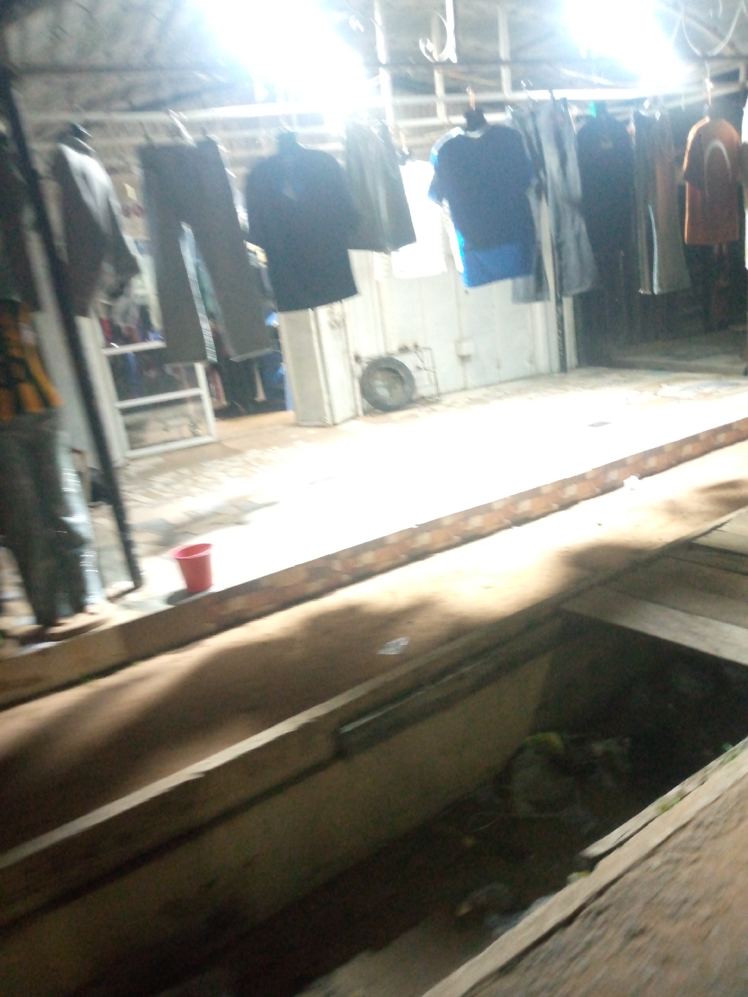 The image depicts a dimly lit indoor market stall in Jalingo, Nigeria, with clothing items hanging on a rack. The stall appears to be open-air or has an open front, with bright overhead lighting creating strong contrasts and shadows. Several garments, including shirts and pants, are visible, some in dark colors like navy blue and black, and one shirt in orange. The stall's floor is made of concrete and wood, with a red bucket placed on the floor near the foreground. In the background, a tire on a stand and some shelving units can be observed. The overall impression is one of a functional, somewhat cluttered, and busy marketplace environment.