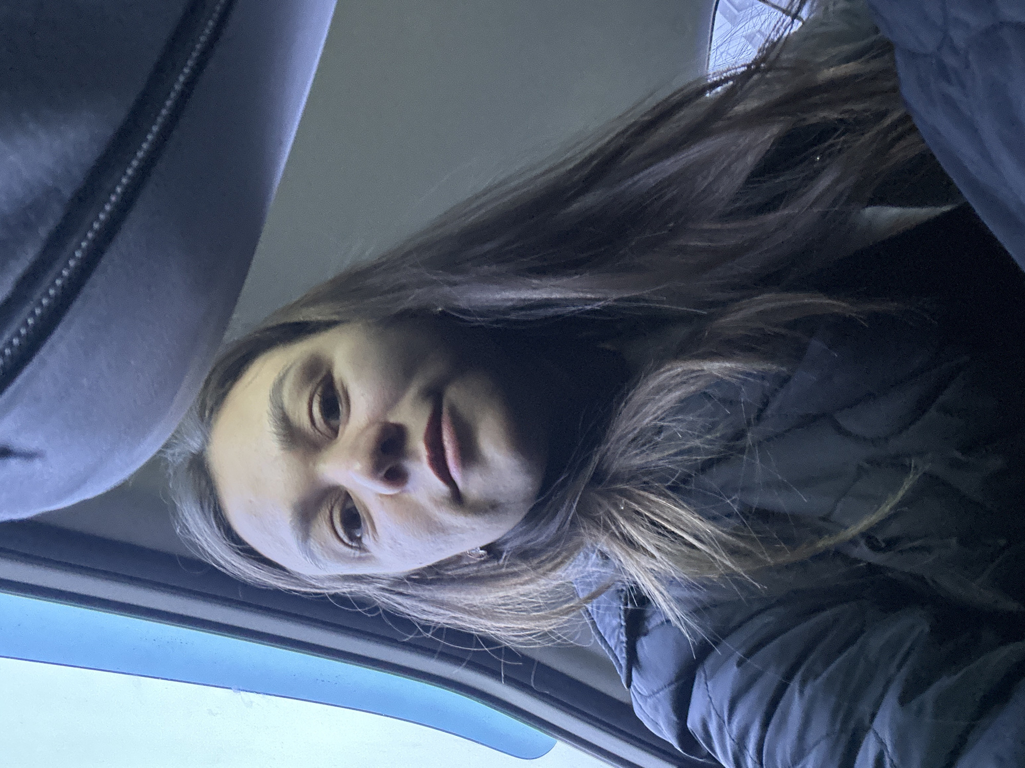 A woman with long brown hair is looking down at the camera. She is wearing a dark quilted jacket. The setting appears to be inside a car, as evidenced by the car roof and window frame visible in the image. The location within the city of Chaikovsky, Russia is not determinable from the image.