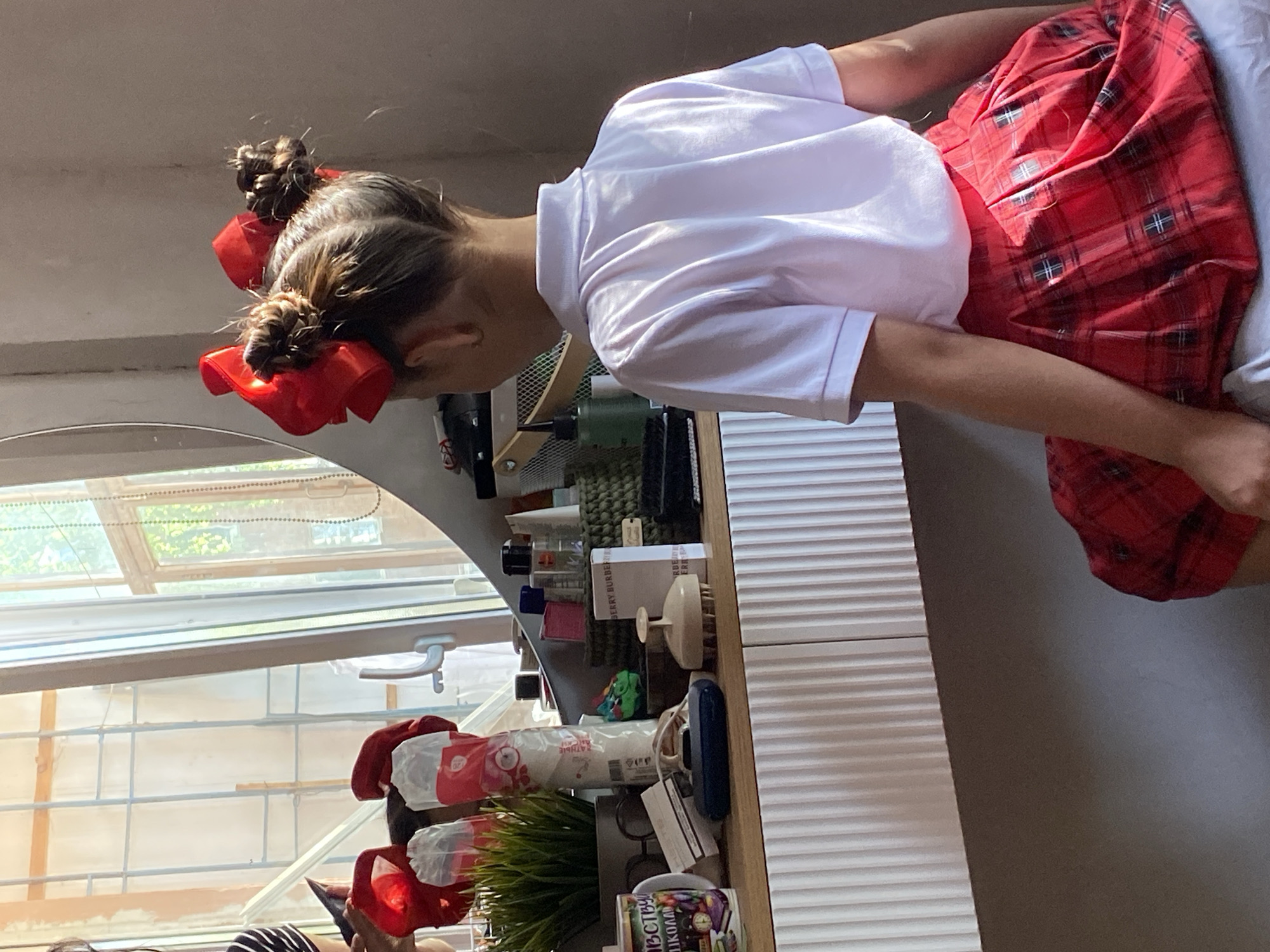 A girl with red bows in her hair stands at a white dresser, wearing a white shirt and red plaid skirt. She appears to be looking at items on the dresser. The scene is indoors, with a window showing an exterior view. The location is Votkinsk, Russia.