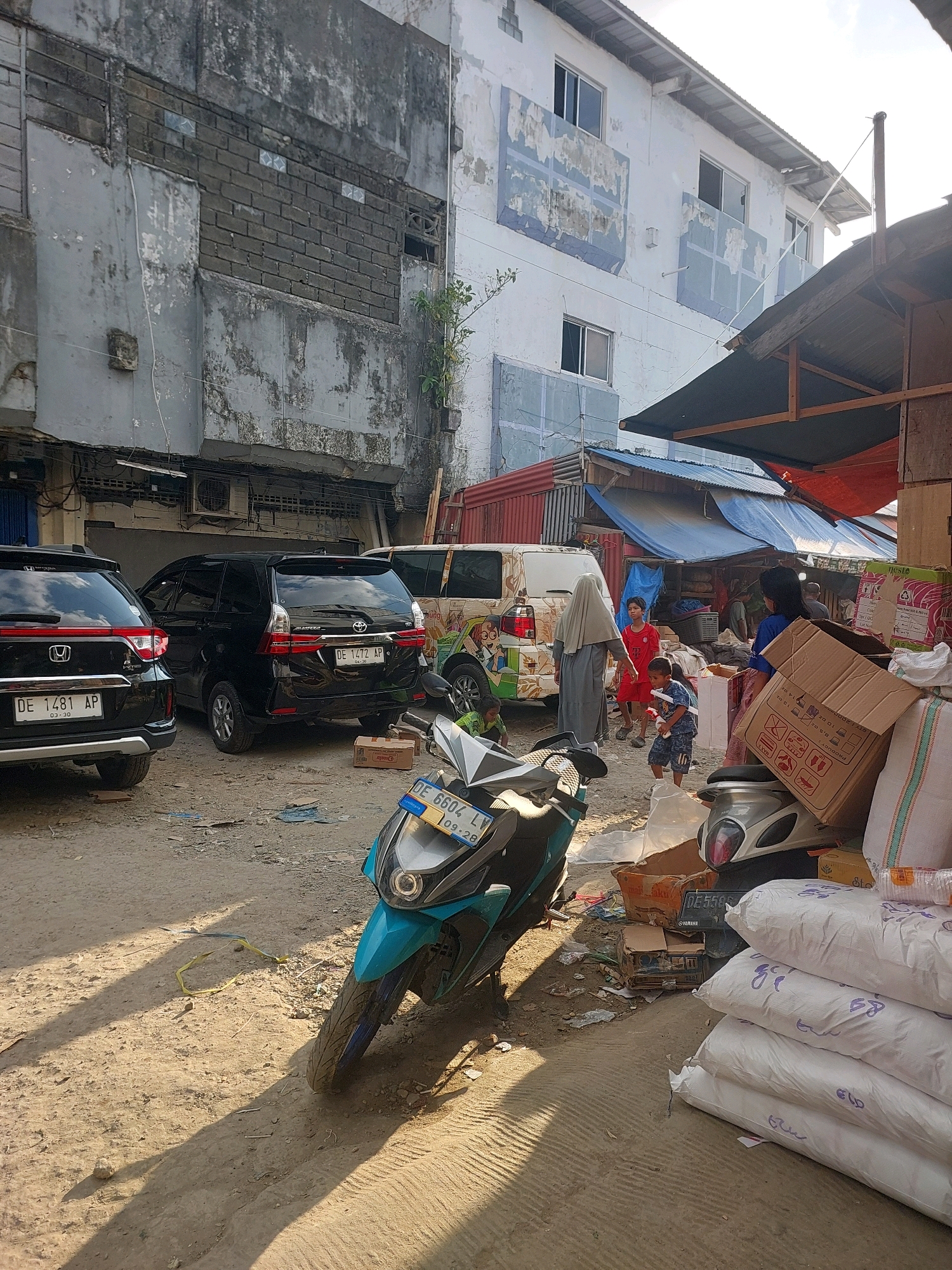 In Ambon, Indonesia, a street scene unfolds with a mix of vehicles, buildings, and people. The foreground is dominated by a teal and silver scooter, casting a long shadow across the dusty, unpaved ground. Behind it, a black SUV is parked, its license plate reading "DE 1472 AP." To its left, the rear of another black car, a Honda with license plate "DE 1481 AP," is visible. Further back, a white van with colorful cartoon decals is parked.
The environment appears to be an urban area with signs of wear and tear. The buildings show weathered facades; one is a multi-story structure with peeling blue paint and visible brickwork, while another, closer to the right, has a corrugated metal roof supported by wooden beams, with tarps providing additional cover.
Several people are present. A woman in a grey hijab walks away from the viewer, facing the white van. A young boy in a red shirt stands near her, looking towards the van, and another young boy in a blue shirt is visible in the middle ground, looking towards the viewer. A woman in a blue shirt is seen interacting with someone behind a stack of large white sacks. Various boxes and bundles are scattered around, including large cardboard boxes and stacks of white sacks, suggesting a commercial or market area. The overall impression is one of a bustling, slightly disorganized, everyday street scene.
