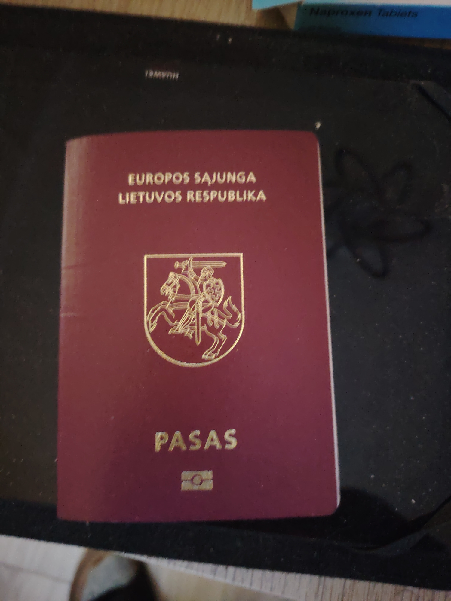 The scene, located in Huntingdonshire, United Kingdom, displays a Lithuanian passport positioned centrally on a dark, textured surface, likely the lid of an electronic device. The passport is burgundy with gold-embossed text and symbols. From top to bottom, the text reads "EUROPOS SAJUNGA," "LIETUVOS RESPUBLIKA," and "PASAS." A gold emblem depicting a knight on horseback (Vytis) is centered below "LIETUVOS RESPUBLIKA." At the bottom, a gold biometric passport symbol is visible. The dark surface exhibits an upside-down "HUAWEI" logo near the upper left quadrant of the passport, indicating the brand of the device. Small, light-colored dust particles are scattered across the dark surface. In the upper right portion of the frame, a partial view of a blue and white box with the text "Naproxen tablets" is present. A subtle, lighter-toned surface is partially visible at the bottom edge of the frame, underlying the dark surface. No individuals are visible within the frame. The ambient lighting suggests an indoor environment.