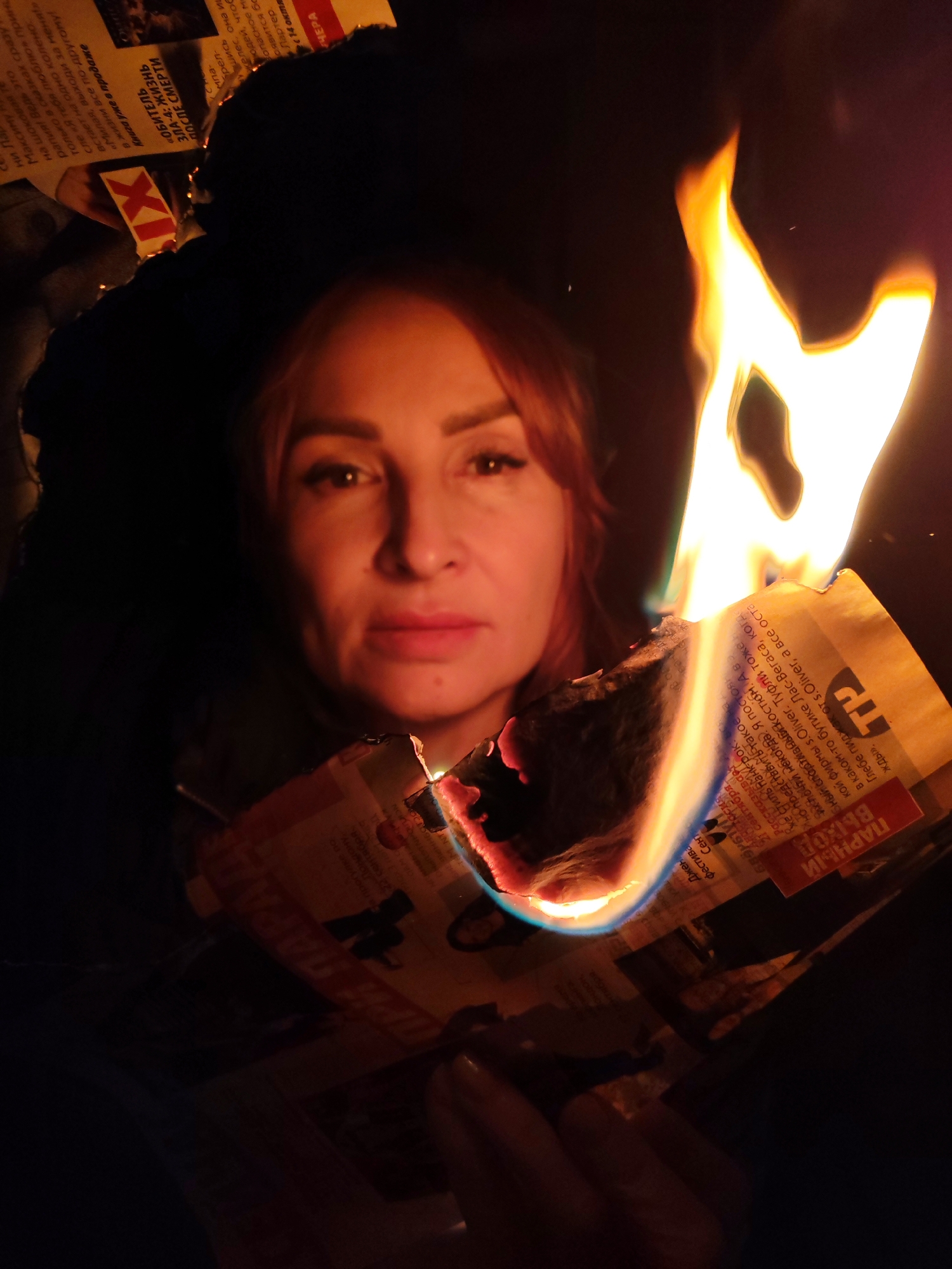 A woman with reddish-brown hair is looking directly at the camera, holding burning newspaper. The newspaper has Russian text, including the words "ПАРНЫЙ ВЫХОД" and references to "s.Oliver" and "Лас-Вегаса." The location is inferred from the OCR text as possibly related to Chaikovsky, Russia, though this is not definitively confirmed within the image itself. The environment is dark, with the primary light source being the fire.