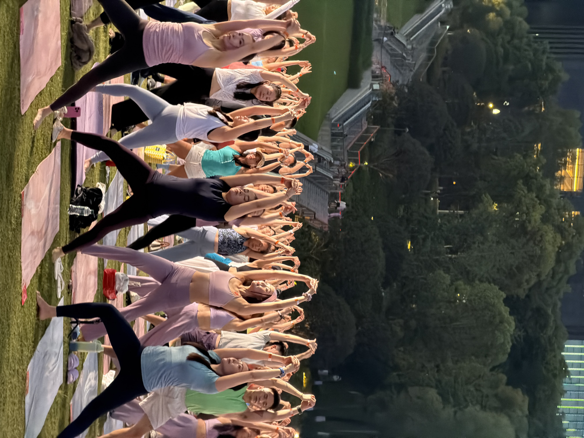 A large group of people, predominantly women, are participating in an outdoor yoga session on a grassy area at dusk. They are arranged in rows on yoga mats, with many holding their arms overhead in a synchronized pose. In the background, there are trees and a multi-tiered stairway with lights. Beyond the trees, city buildings are visible, including one with illuminated windows. The overall scene suggests a community event or class held in a park setting within an urban environment.