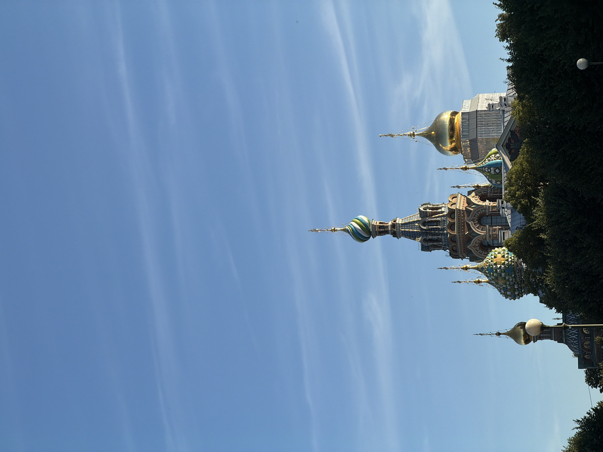The image captures a detailed view of the Church of the Savior on Spilled Blood, a prominent landmark in Saint Petersburg, Russia. The church's distinct onion domes, adorned with vibrant mosaic patterns and golden spires, are silhouetted against a clear blue sky streaked with wispy clouds. The architectural details, including intricate facades and ornate finials, are visible, highlighting the grandeur of the structure. Lush green trees form a dense foreground, partially obscuring the lower portion of the building. The scene is illuminated by natural daylight, suggesting it was taken during the day. No people or specific actions are depicted.