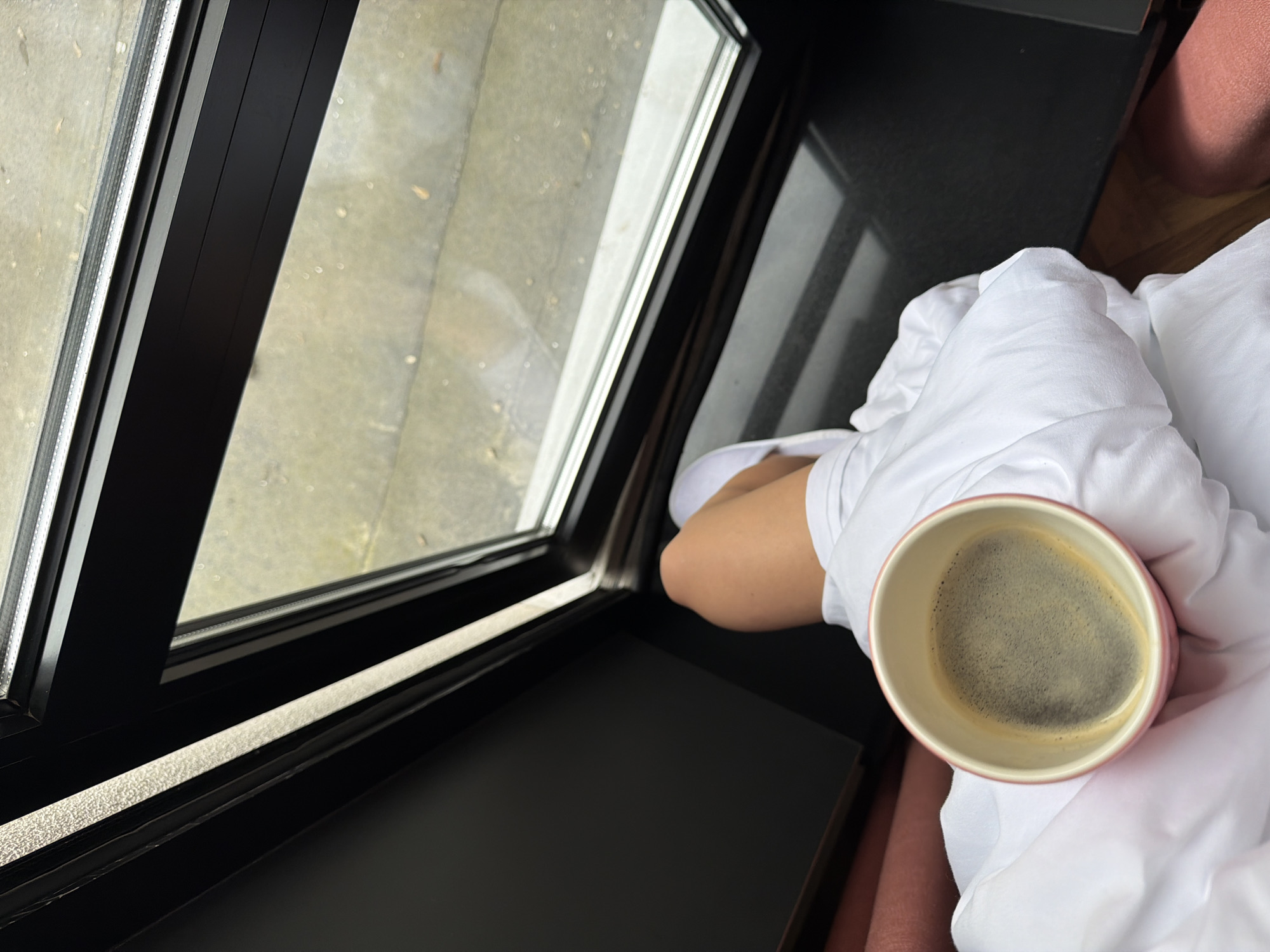 A high-angle shot shows a person holding a pink mug of coffee while sitting near a window. The window has a black frame and shows an outside view of a gray concrete surface. The person is covered in a white blanket or sheet. A black surface, possibly a table, is visible next to the window. Part of a pink upholstered surface is visible on the right edge of the image. There is no indication of a specific city or location.