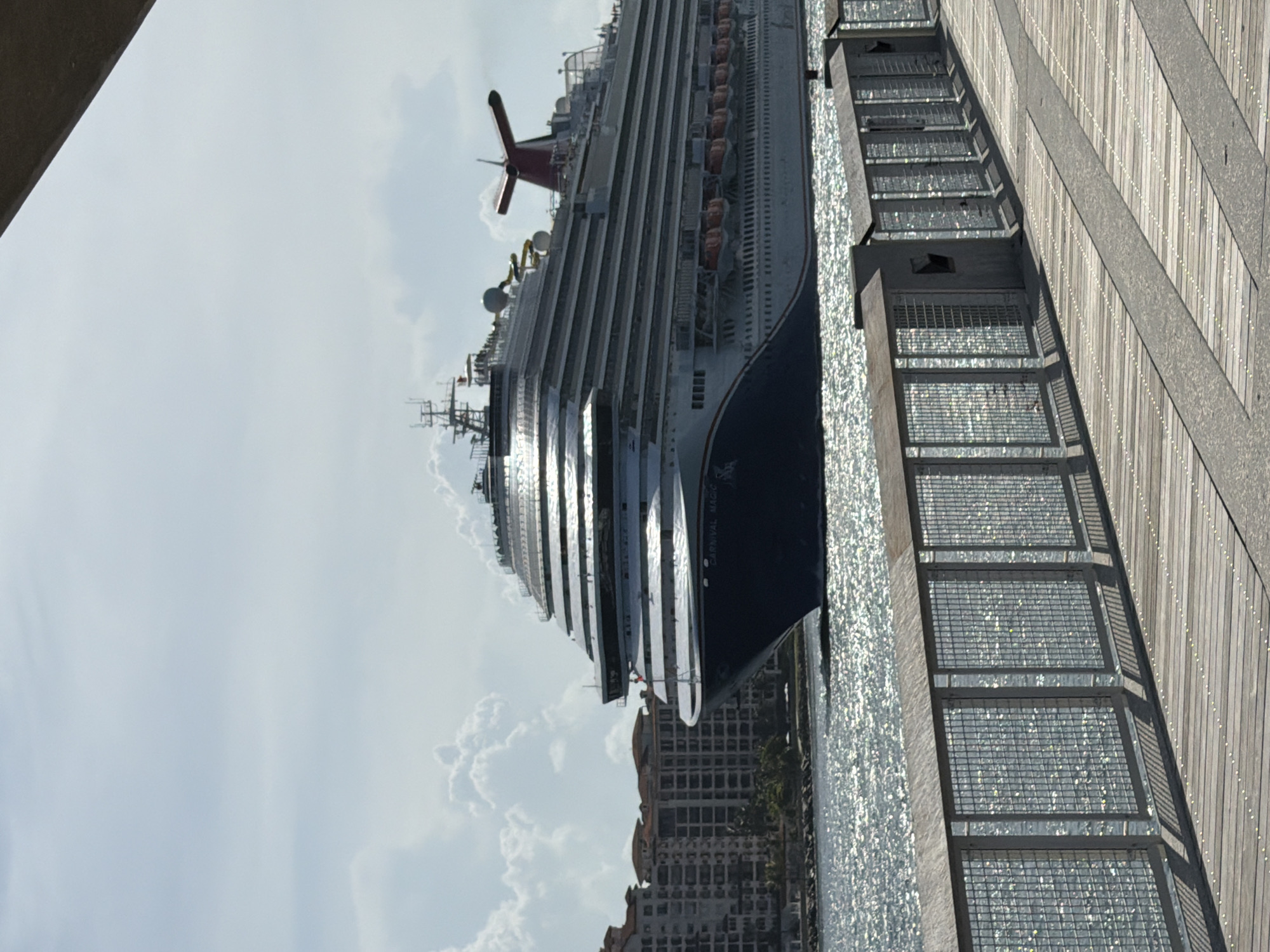 A large cruise ship, identified by the name "Carnival Magic" on its hull, is seen from the side, sailing on a bright, sunny day. The ship is predominantly white with a dark blue hull. Its upper decks feature multiple levels of balconies and windows, and a distinctive red funnel with a white logo is visible at the stern. Lifeboats are clearly mounted along the sides of the ship.
In the background, a row of multi-story buildings with red-tiled roofs lines the shore. The water in front of the ship and the buildings is a sparkling blue, reflecting the sunlight. In the foreground, a wooden boardwalk with metal railings runs along the edge of the water. The boardwalk is made of light-colored wooden planks and the metal railings are made of a mesh material. Shadows from the railings are cast onto the boardwalk. The sky is a pale blue with scattered white clouds. The scene appears to be a waterfront area in Miami Beach, United States of America, likely a port or promenade, given the presence of the cruise ship and the boardwalk. No people are clearly discernible on the ship or the boardwalk, though there are indications of people on the upper decks of the ship. The overall impression is one of a busy maritime environment on a clear day.