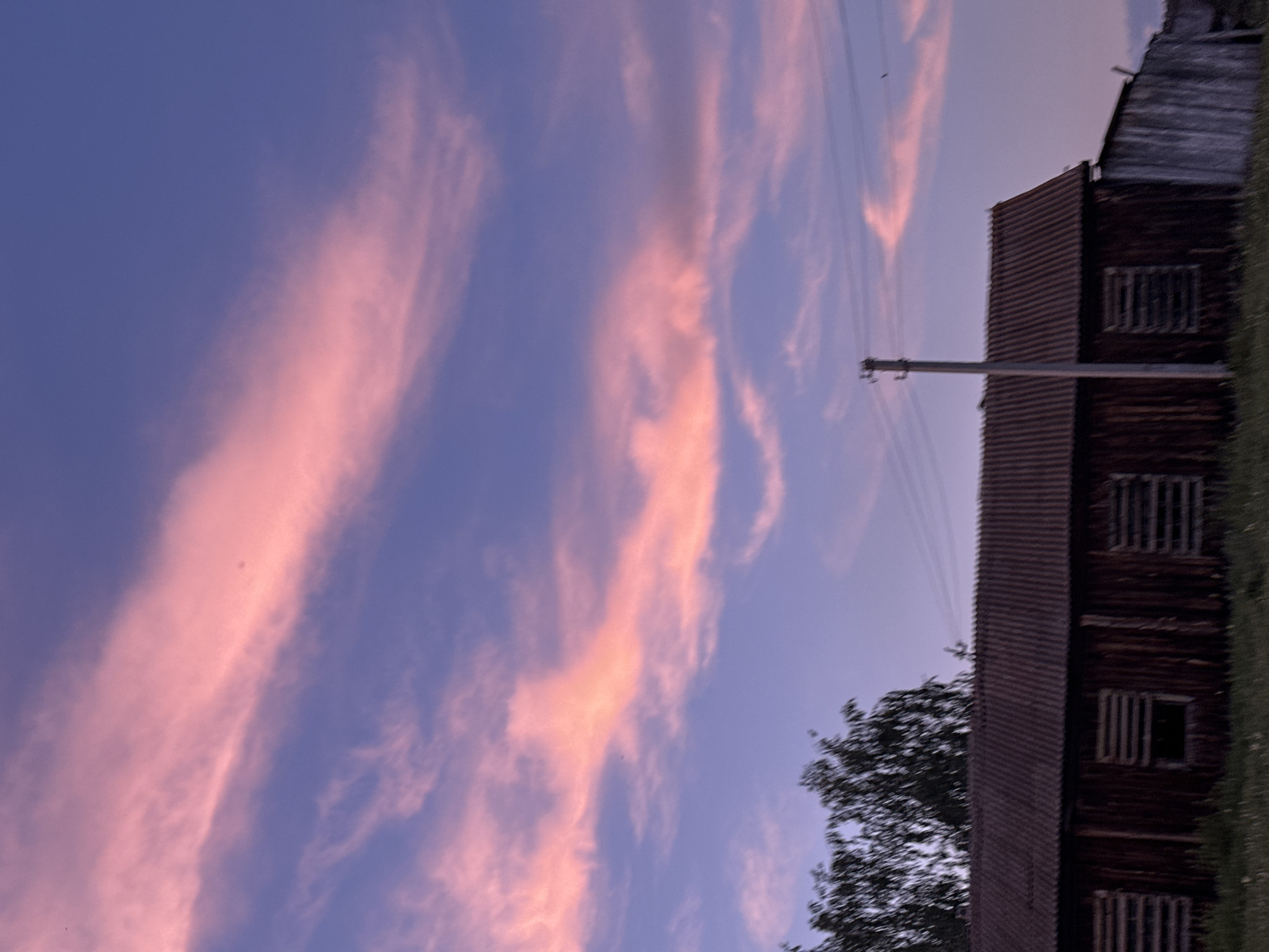 An outdoor, eye-level shot depicts a rustic, wooden building with a corrugated roof under a twilight sky. The sky is a gradient of deep blue to a soft lavender, streaked with wispy, pink-tinged clouds. A single wooden utility pole stands to the right of the building, with power lines stretching across the upper portion of the frame. The building itself appears aged, with vertical wooden planks and three boarded-up window openings on its visible facade. To the left of the building, a dense cluster of dark green trees rises against the colorful sky. The foreground consists of a grassy area, with the lower part of the building slightly obscured by its dark shadow. The overall atmosphere is serene and rural.