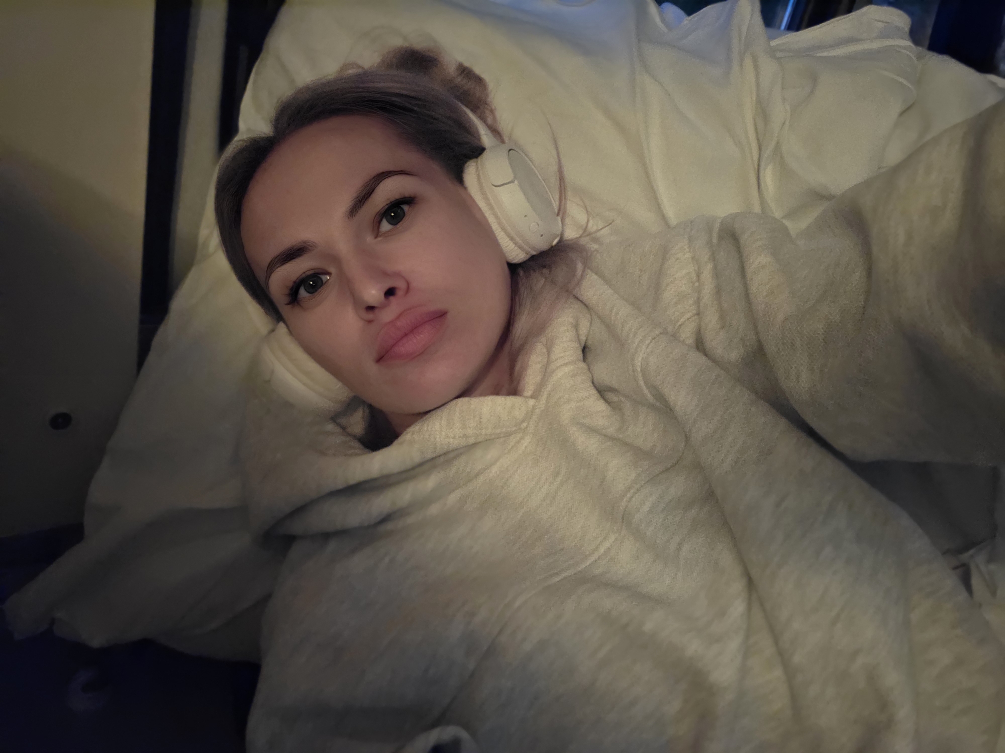A woman is lying on a bed, wearing a light gray hooded sweatshirt and white headphones around her neck. She has blonde hair pulled back and is looking directly at the camera with her lips slightly parted. The bed is covered with white linens. The scene appears to be indoors, possibly a bedroom, with dark surroundings and hints of warm lighting. There is no indication of the location being in the city of Korshunikha, Russia, or any other specific geographical place.