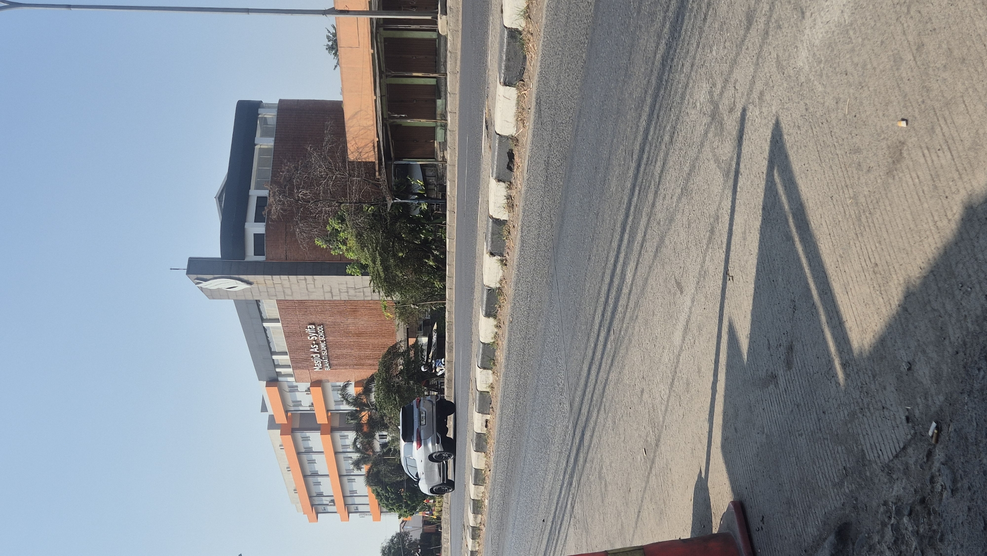 The image shows the As-Syifa Islamic School in Karawang, Indonesia, on a clear, sunny day. The modern building features a prominent tower with a stylized leaf logo and a facade of textured orange brick. The building is multi-story with orange and white accents. In the foreground, a silver SUV is driving on a road that separates the school from a row of shops with closed shutters. The road is lined with concrete barriers. The ground in the lower foreground is a concrete surface with striped patterns and a traffic cone. Shadows are cast across the ground from the sun. There are no visible people interacting directly with the building or each other. The environment appears to be an urban or semi-urban setting with a clear blue sky.