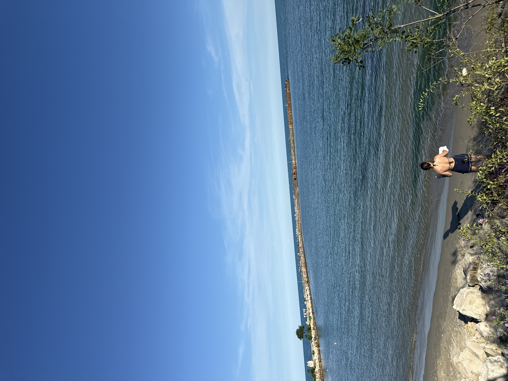 A lone woman stands on a sandy beach, facing the calm, blue water. She is wearing a dark bikini top and dark shorts, and is holding a white towel in her right hand. The beach is lined with large, light-colored rocks, and green foliage with purple flowers grows along the edge of the sand. In the distance, a long pier extends out into the water, with several people visible standing on it. The sky above is a clear, bright blue, with a few wispy white clouds. The scene appears peaceful and serene. The image was taken at an unknown location in an unknown city.