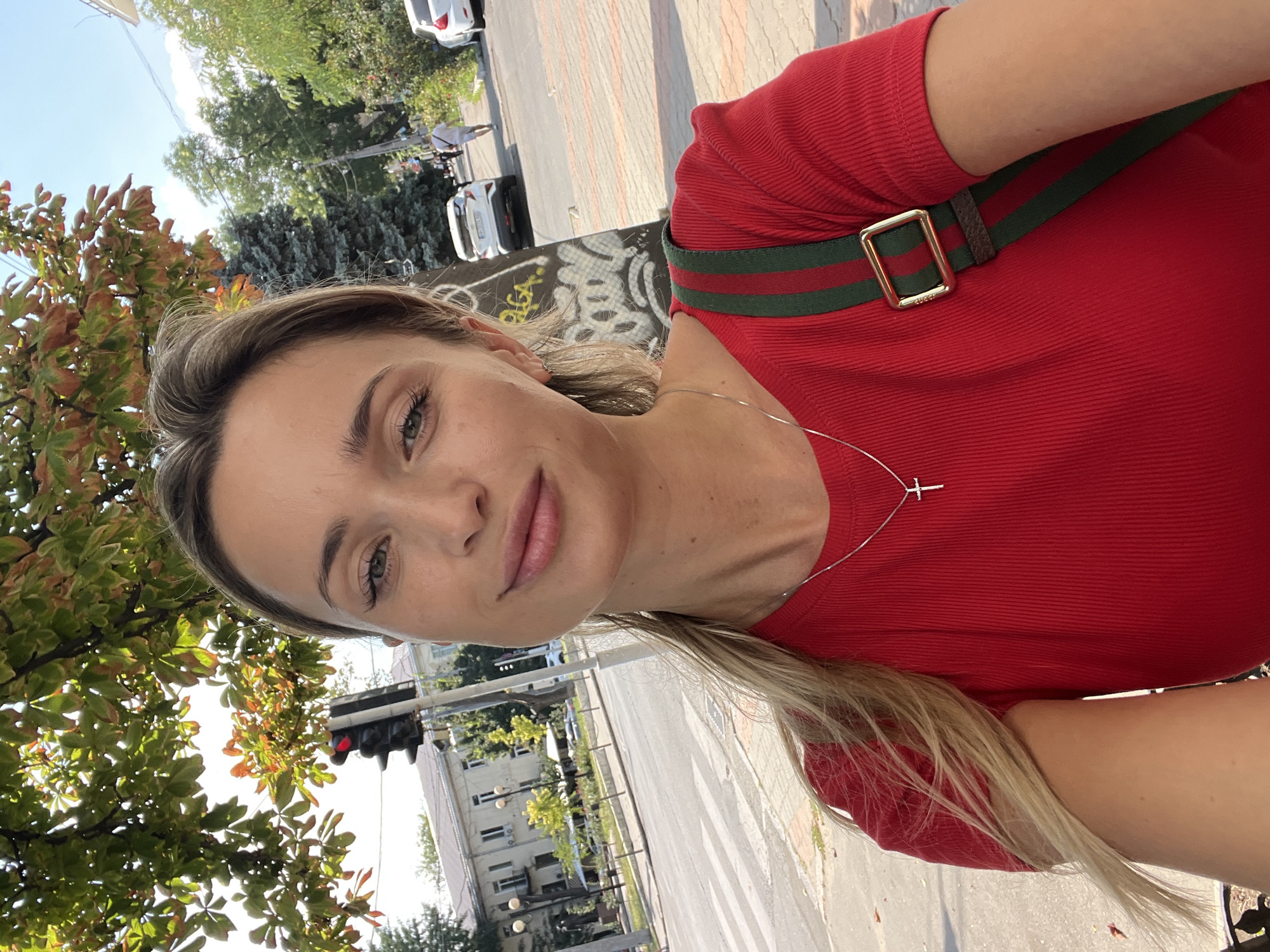 A medium shot taken from a slightly low angle captures a blonde woman in her late 20s or early 30s standing on a sidewalk in Odesa, Ukraine. She is looking directly at the camera, her gaze steady and her lips curved into a slight smile. The woman is wearing a red ribbed shirt and a silver necklace with a cross pendant. A bag with green and red straps is slung over her right shoulder.
The background reveals a city street scene under a bright, clear sky. To her left, a traffic light shows a red signal, and a leafy tree with some autumnal colored leaves hangs over the frame. Across the street, there are buildings, lampposts, and a pedestrian crossing. Several cars are visible on the road, and a man in a light-colored shirt and shorts is walking away from the camera down the sidewalk on the right. Graffiti is present on a pillar behind the woman. The overall impression is of a casual, sunny day in an urban environment.