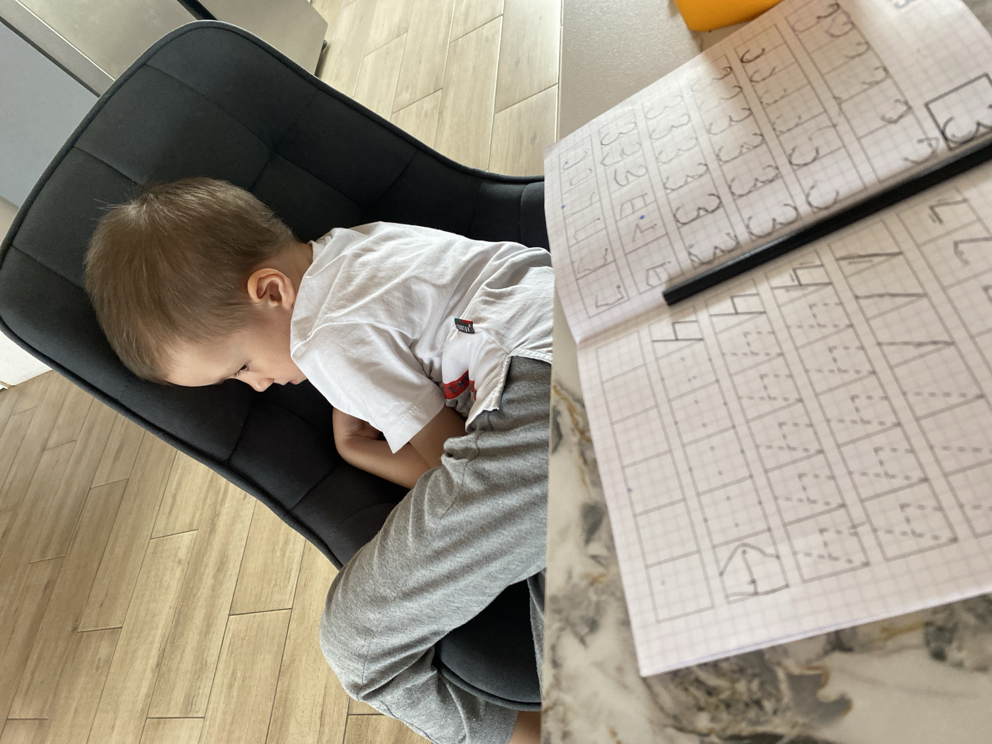 A young boy with fair hair and a white T-shirt is sitting in a dark gray chair in what appears to be a home setting. His head is resting on the back of the chair, suggesting he may be resting or sleeping. To the right of the boy is a sheet of graph paper with handwritten letters, possibly Cyrillic, indicating a learning activity. A black pencil is placed on the paper. The floor is made of light-colored wooden planks. The picture's location is likely an indoor domestic environment, possibly in Chaikovsky, Russia, given the prompt.
