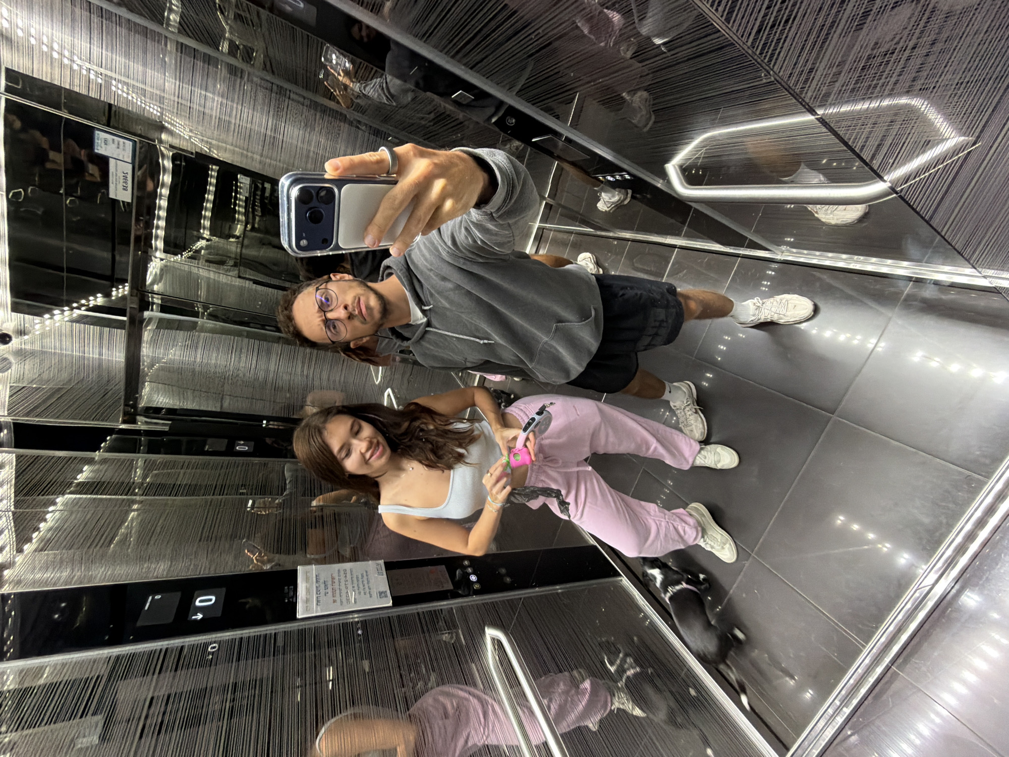 A man and woman are taking a selfie in an elevator. The man holds a phone, wearing glasses, a grey hoodie, and black shorts. The woman is beside him, wearing a white tank top and pink sweatpants, holding a pink object. A small black-and-white dog is also in the elevator. The elevator interior features reflective walls and ceiling. The elevator panel shows floor "0". The elevator is in Tel Aviv, Israel, based on the Hebrew writing seen on the panel.